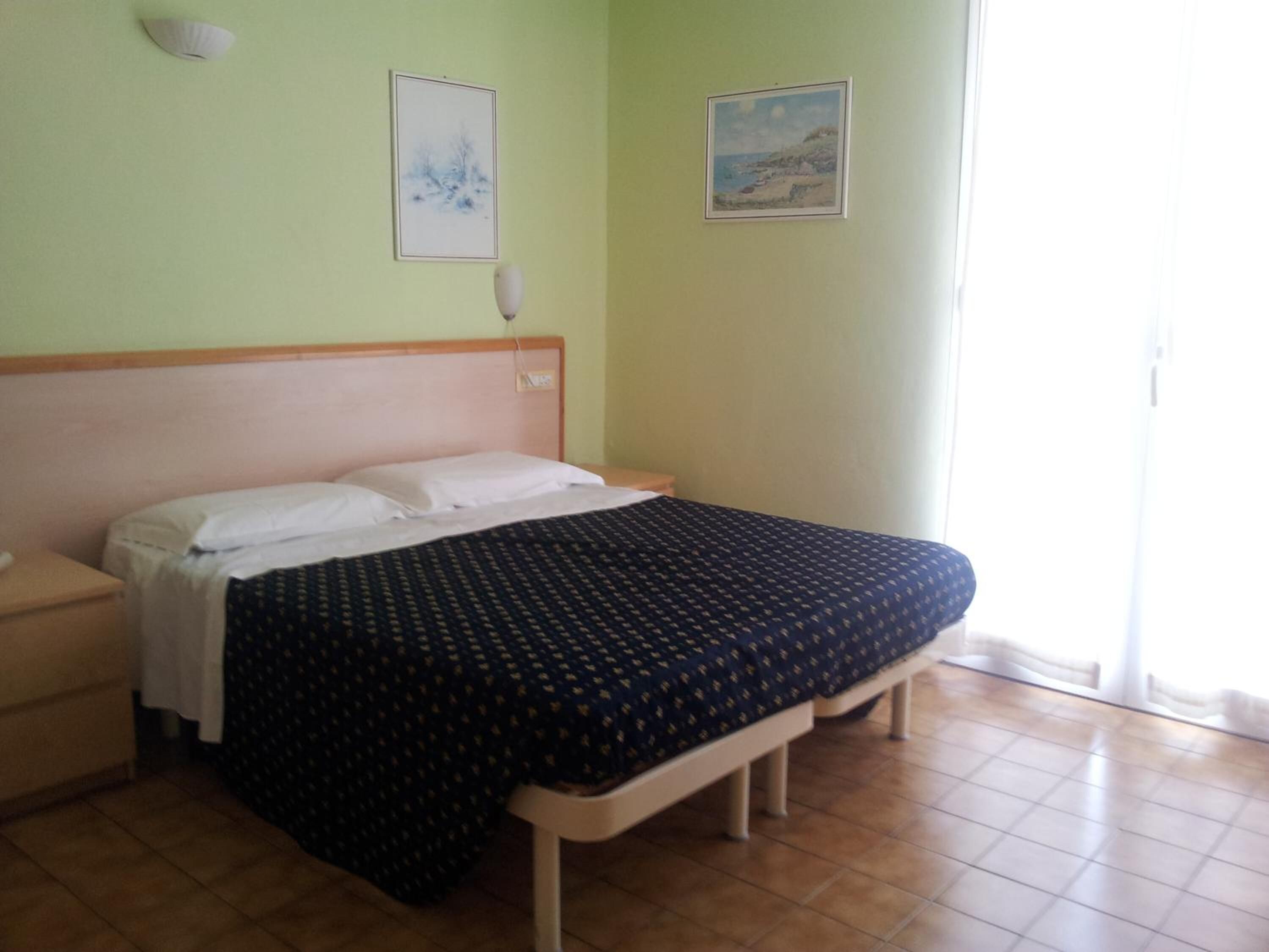 Un dormitorio con cama, mesita de noche, lámpara de pared y dos cuadros enmarcados.
