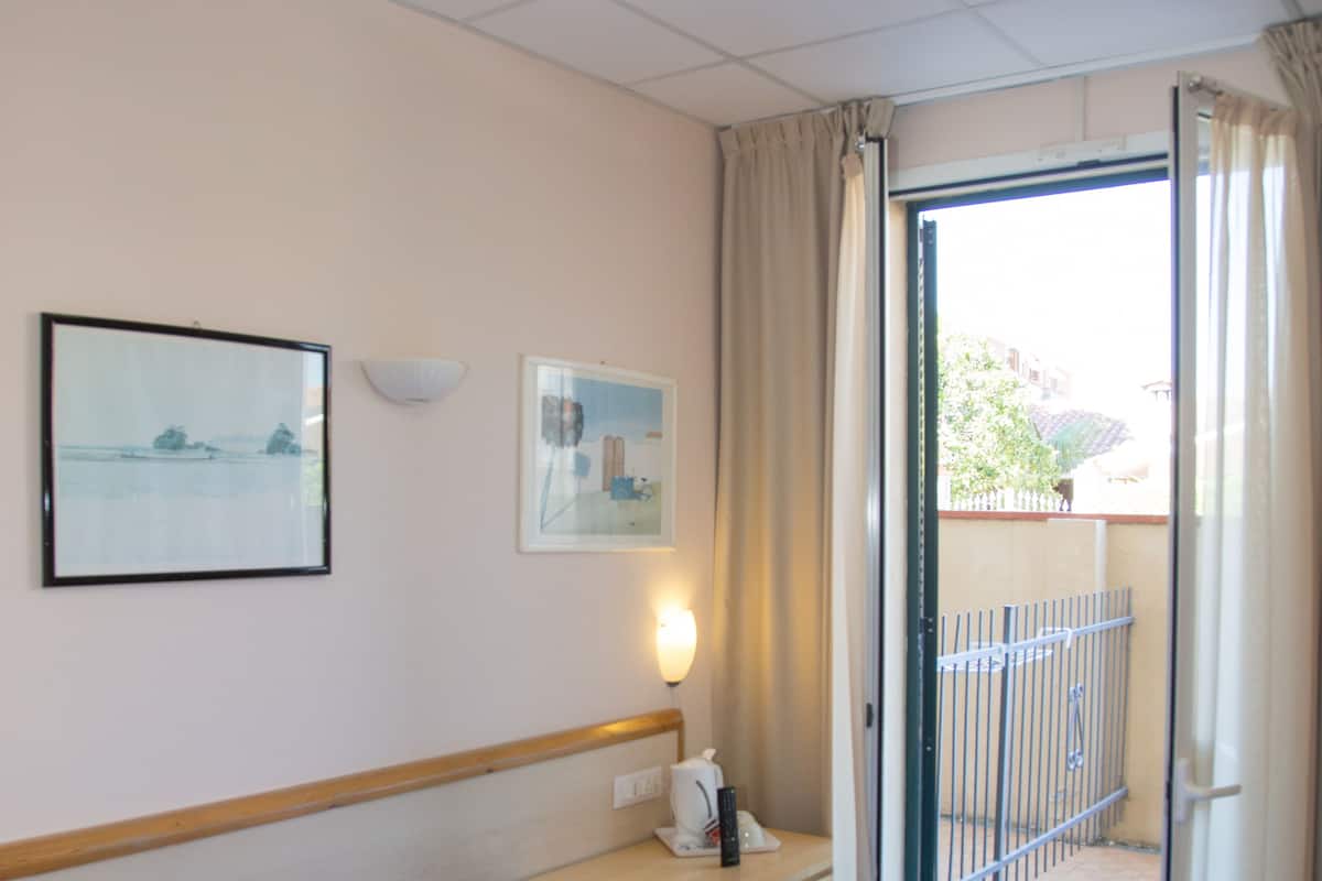 A hotel room with two beds, a nightstand, a lamp, and a view of the outside.