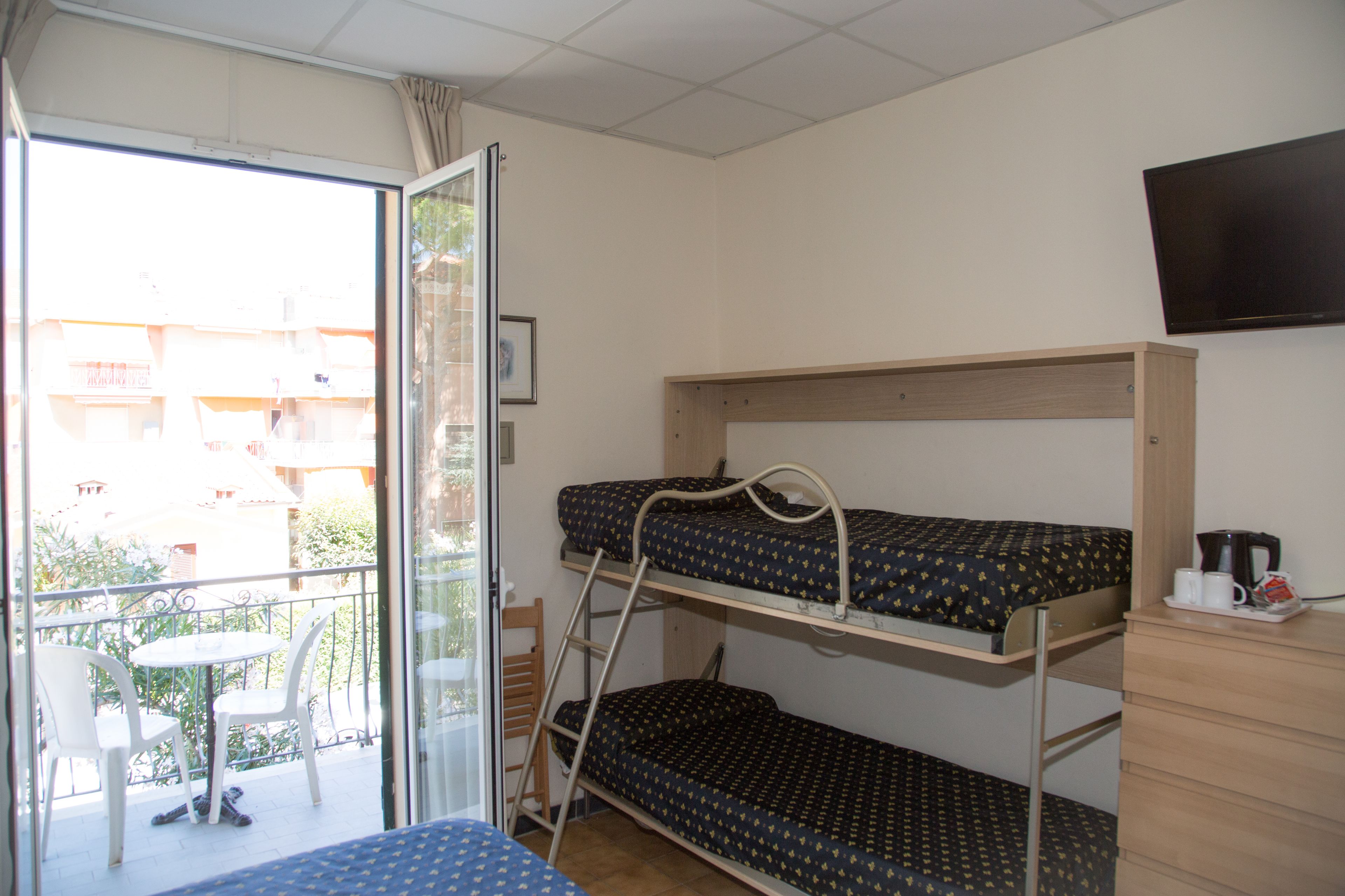 Habitación tipo dormitorio con literas, una pequeña mesa, sillas, una escalera, un televisor y un balcón con vistas a edificios.