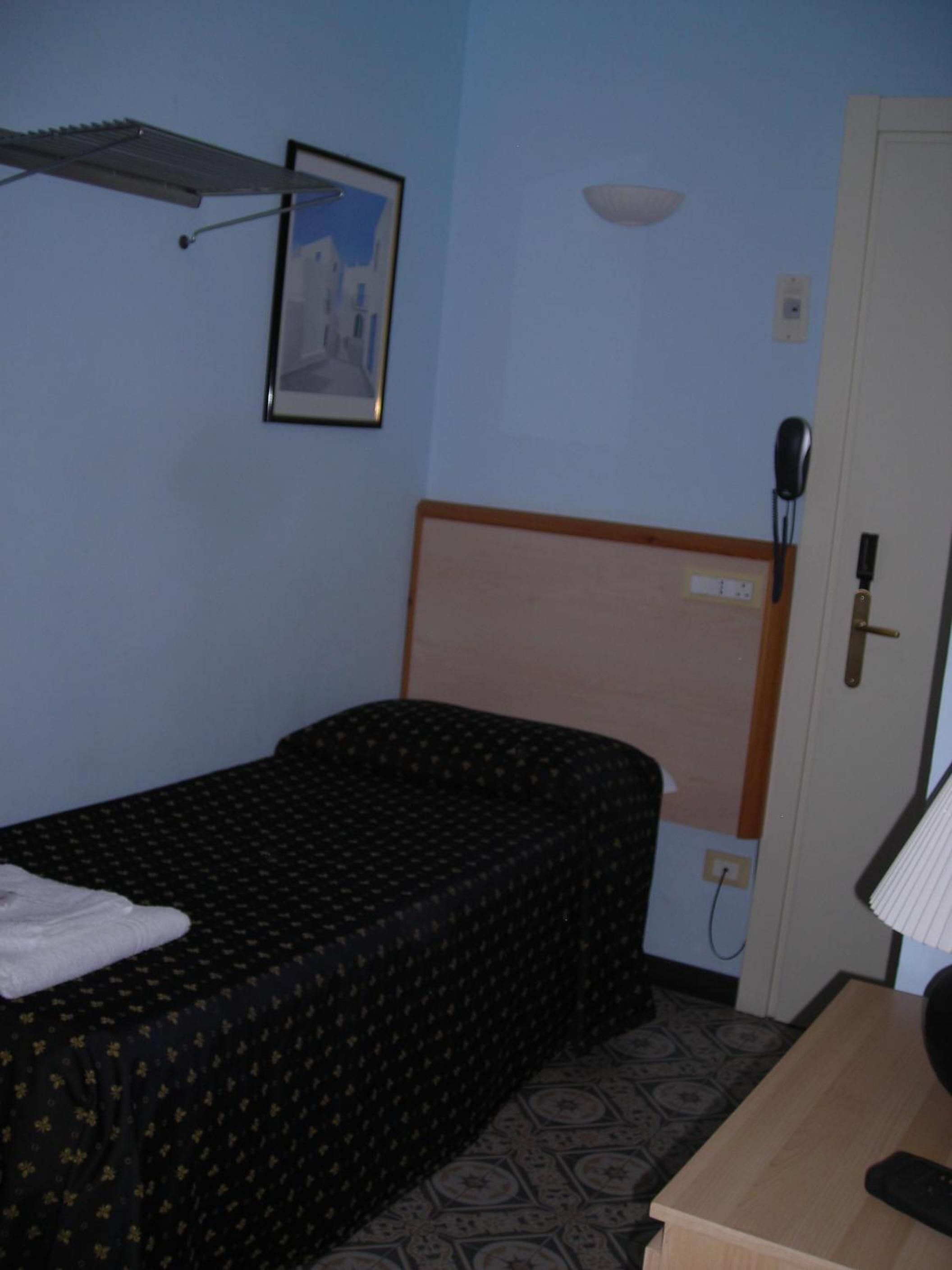 Una piccola camera con un letto singolo, una testiera in legno, un comodino con lampada, un telefono e un quadro appeso al muro.