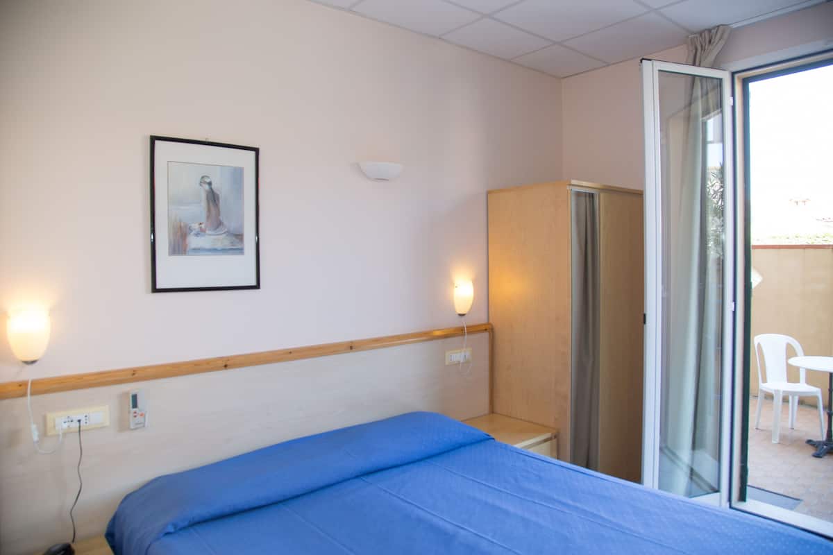 A hotel room with a bed, a framed picture on the wall, a wardrobe, and a sliding glass door leading to an outdoor area with a chair.