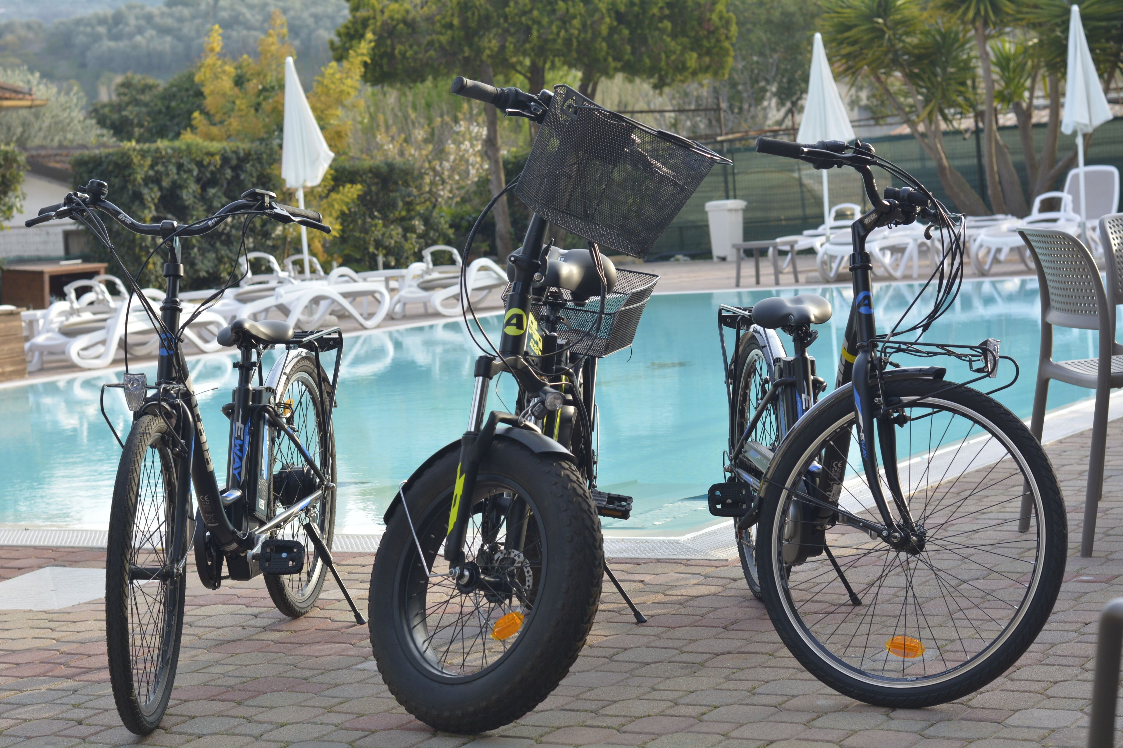 Tre biciclette parcheggiate vicino a una piscina, con lettini e ombrelloni sullo sfondo.