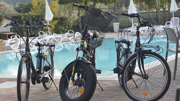 Tre biciclette parcheggiate vicino a una piscina, con lettini e ombrelloni sullo sfondo.