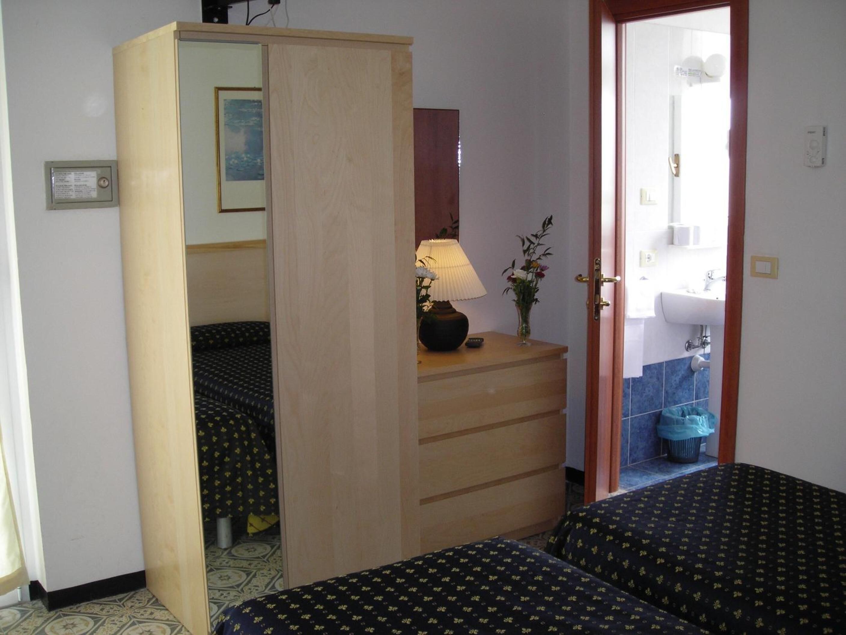 Un dormitorio con dos camas, un armario de madera, una mesita de noche con lámpara y un baño visible a través de una puerta abierta.