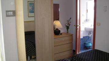 Un dormitorio con dos camas, un armario de madera, una mesita de noche con lámpara y un baño visible a través de una puerta abierta.