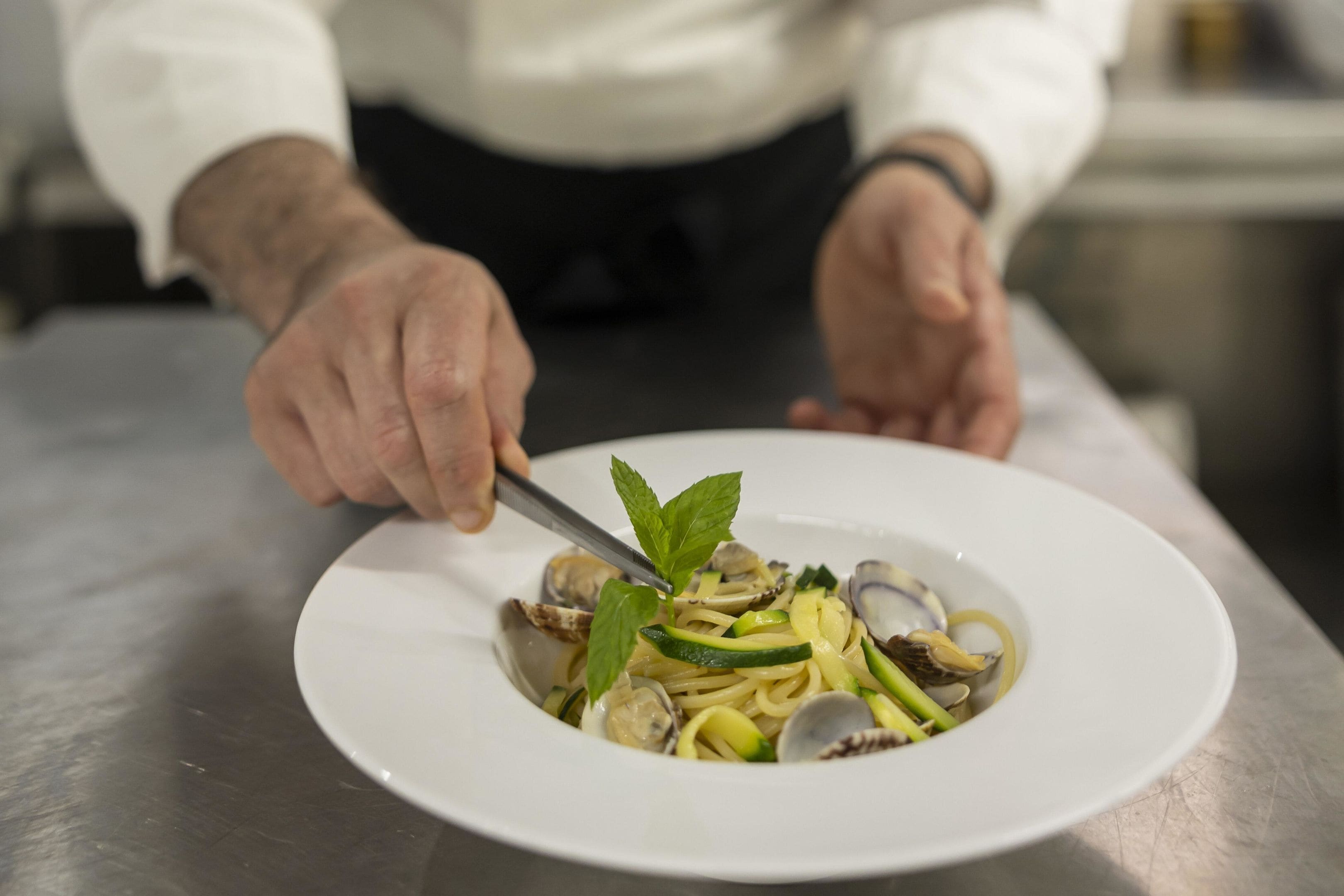 A chef's hands holding a fork over a plate of pasta with clams and vegetables.