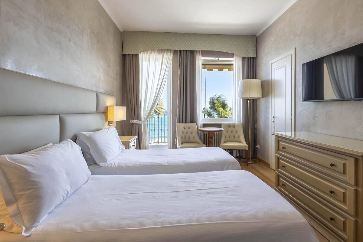 Ein Hotelzimmer mit zwei Betten, einem Fernseher, einem Schreibtisch und einem Balkon mit Meerblick.
