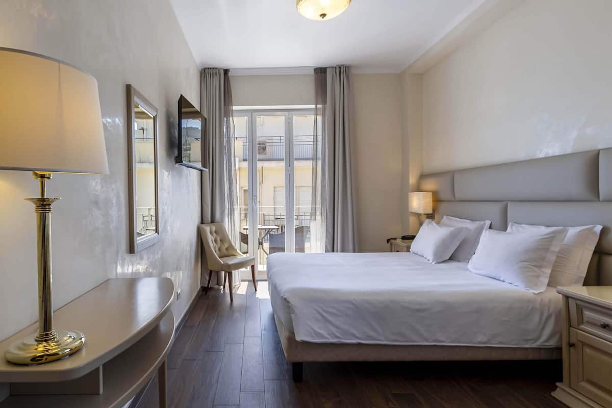Ein Hotelzimmer mit einem großen Bett, Nachttischen, einem Schreibtisch, einem Sessel und einem Fenster mit Vorhängen.