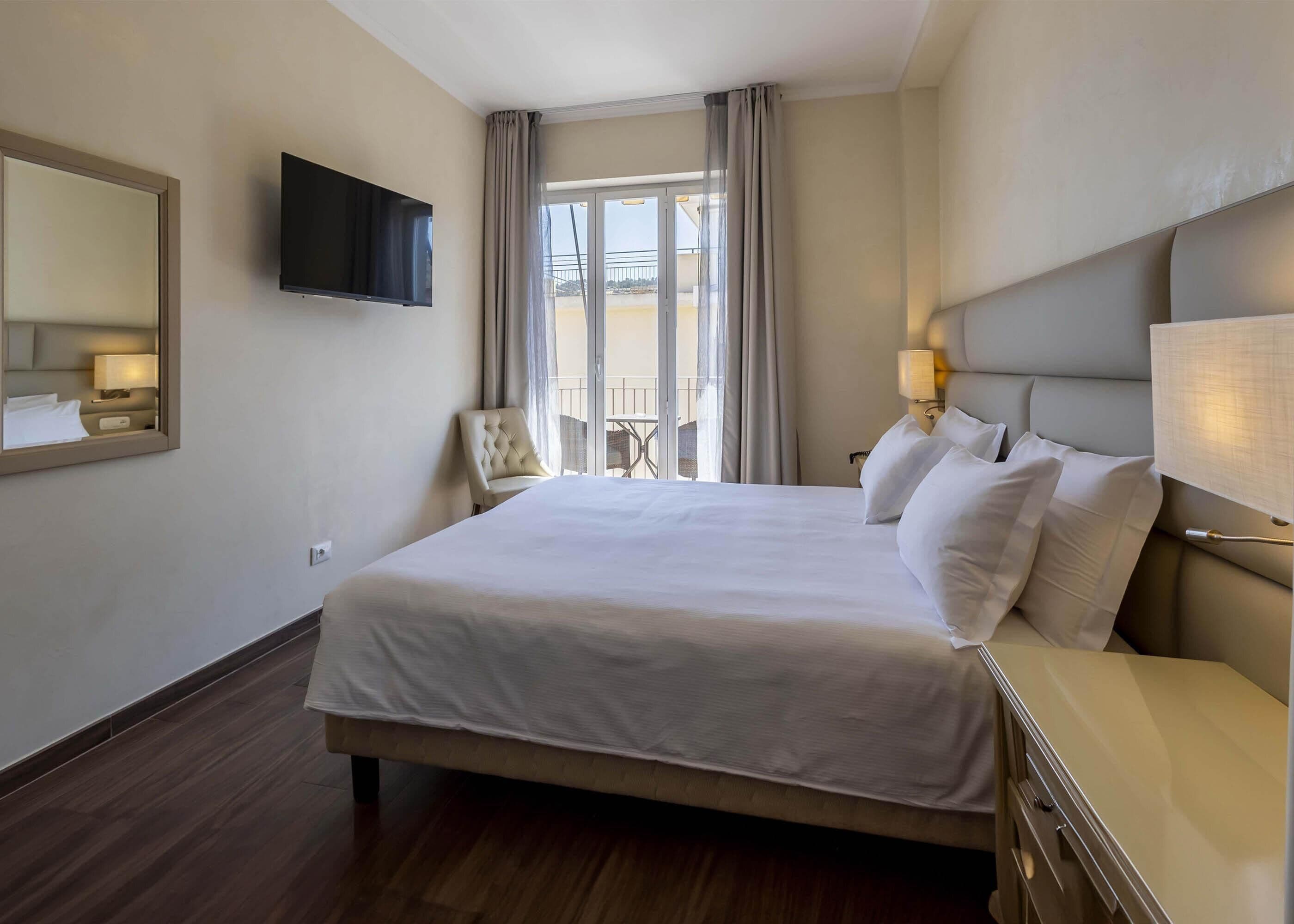 Habitación de hotel con una cama grande, un televisor de pantalla plana montado en la pared, una silla, una mesita y un balcón con vistas.