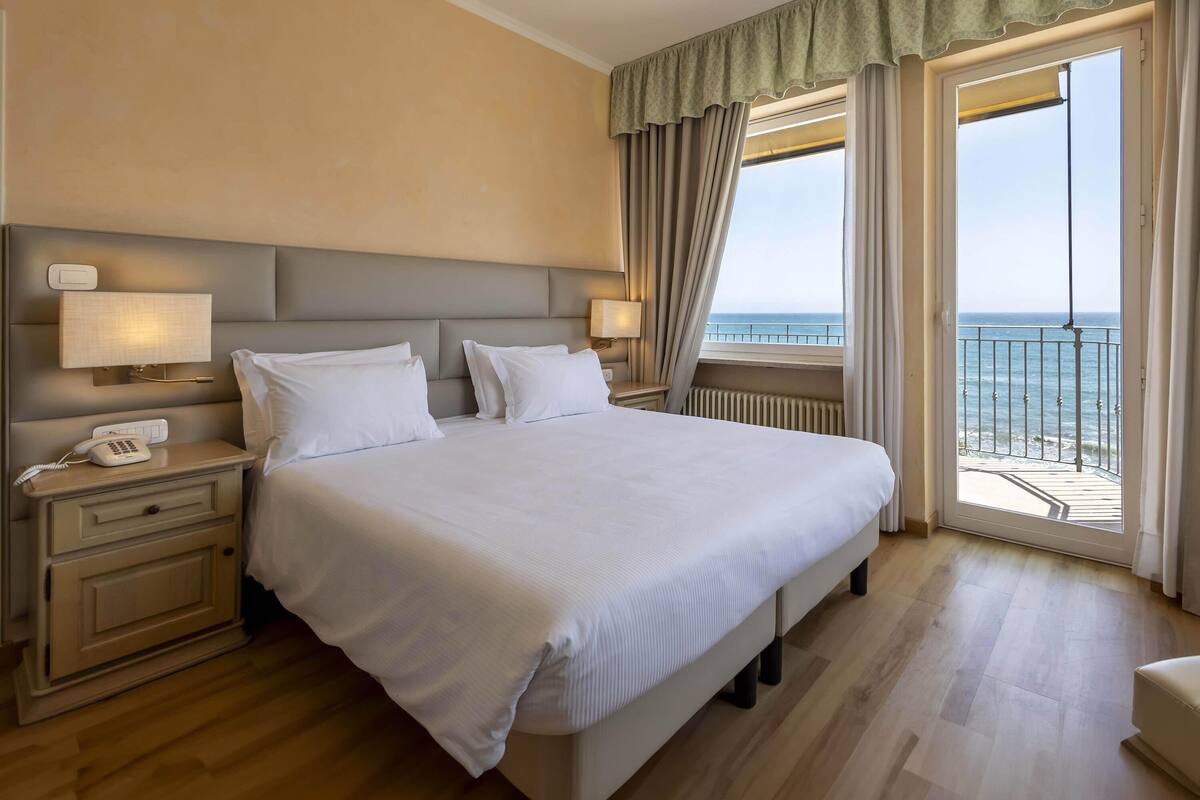 Ein Hotelzimmer mit einem großen Bett, Nachttischlampen, einem Telefon auf dem Nachttisch und Meerblick vom Balkon.