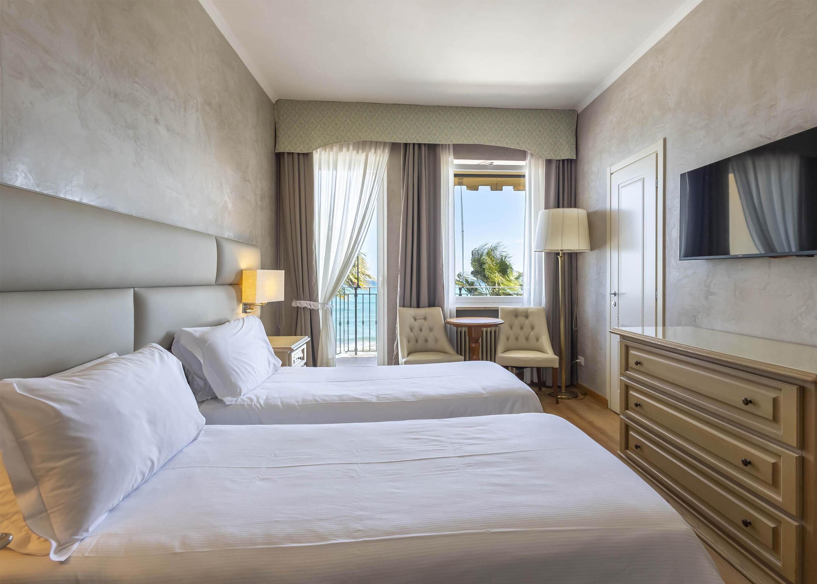 A hotel room with two beds, a TV, a desk, and a balcony with a view of the sea.