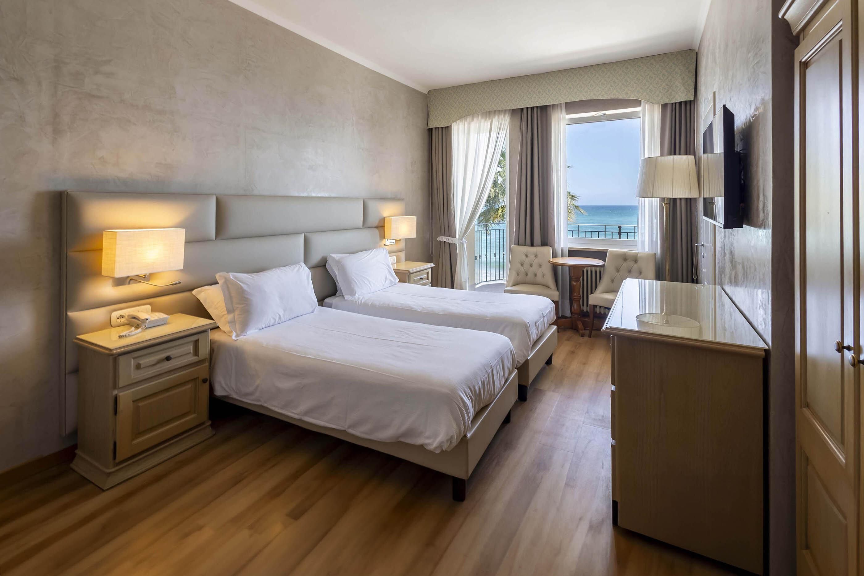A hotel room with two beds, a desk, a chair, a TV, and a balcony with a view of the sea.