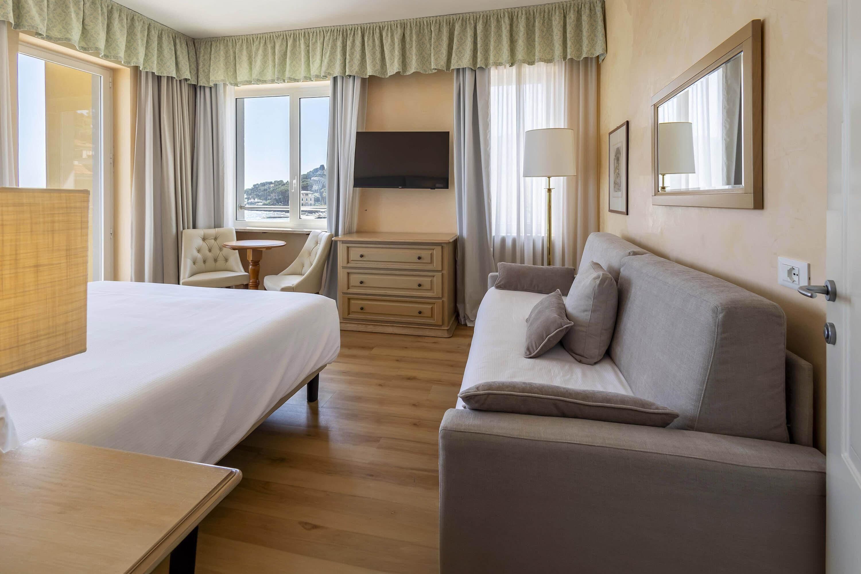 Habitación de hotel con cama, sofá, escritorio, silla, televisión y ventana con cortinas.