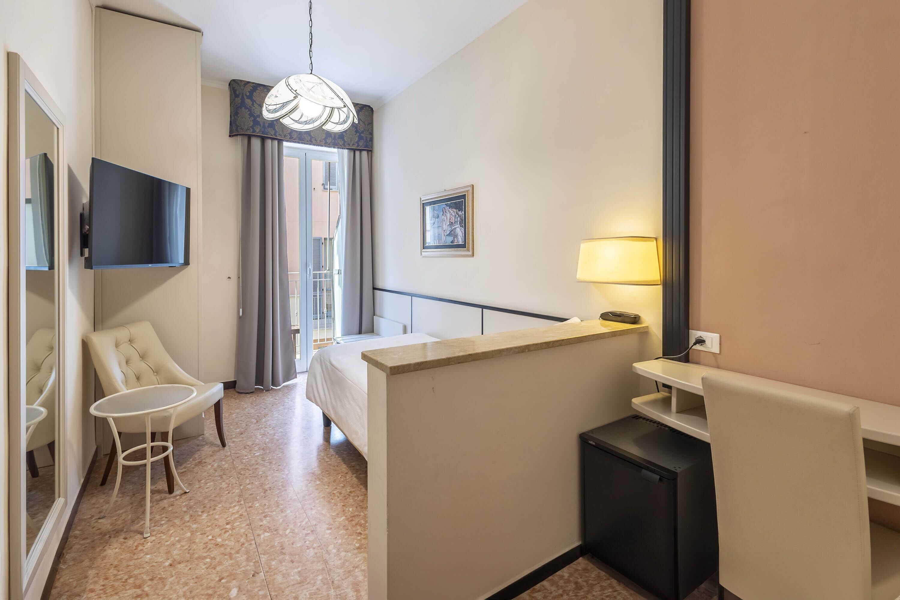 A hotel room with a bed, a desk, a chair, a small table, a TV mounted on the wall, a lamp, and a picture on the wall.
