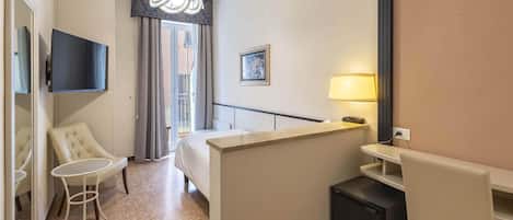 A hotel room with a bed, a desk, a chair, a small table, a TV mounted on the wall, a lamp, and a picture on the wall.