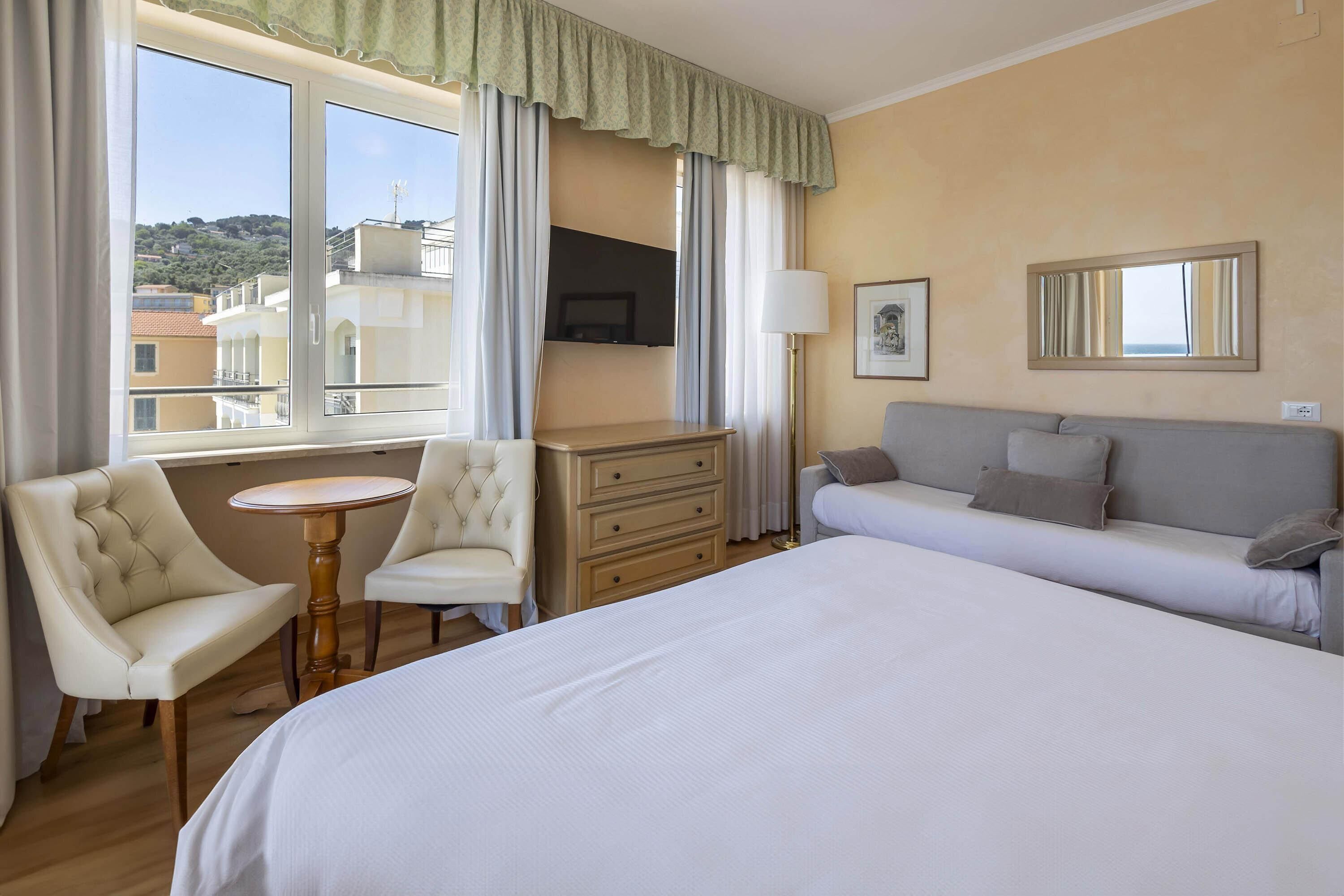 A hotel room with a bed, a sofa, a desk with a chair, a TV mounted on the wall, and a window with curtains.