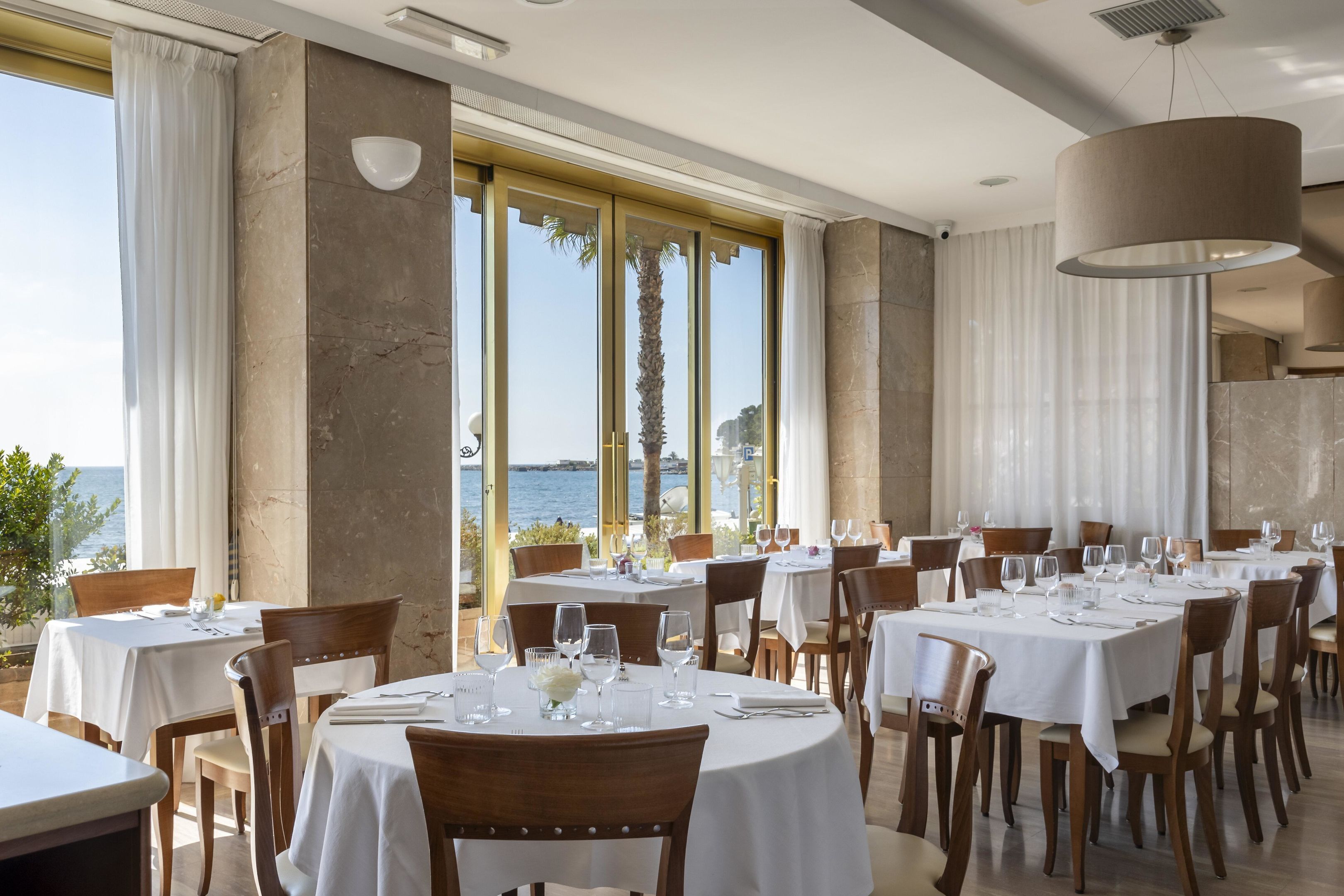 Un restaurant avec des tables rondes, des nappes blanches et des chaises en bois. De grandes fenêtres offrent une vue sur la mer.