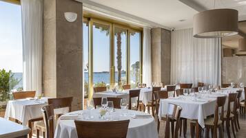 Un restaurant avec des tables rondes, des nappes blanches et des chaises en bois. De grandes fenêtres offrent une vue sur la mer.
