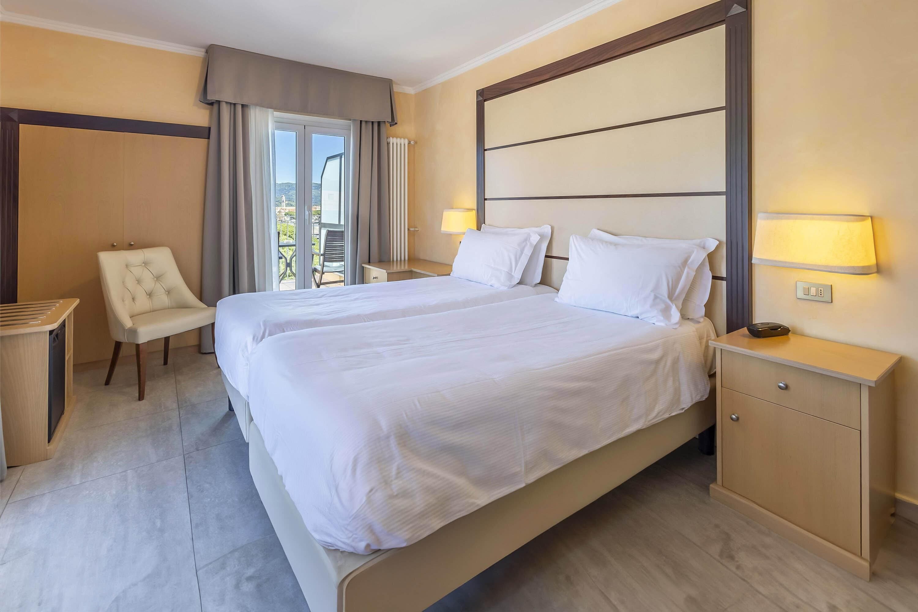 A hotel room with a large bed, a chair, a bedside table, and a view of the outdoors.