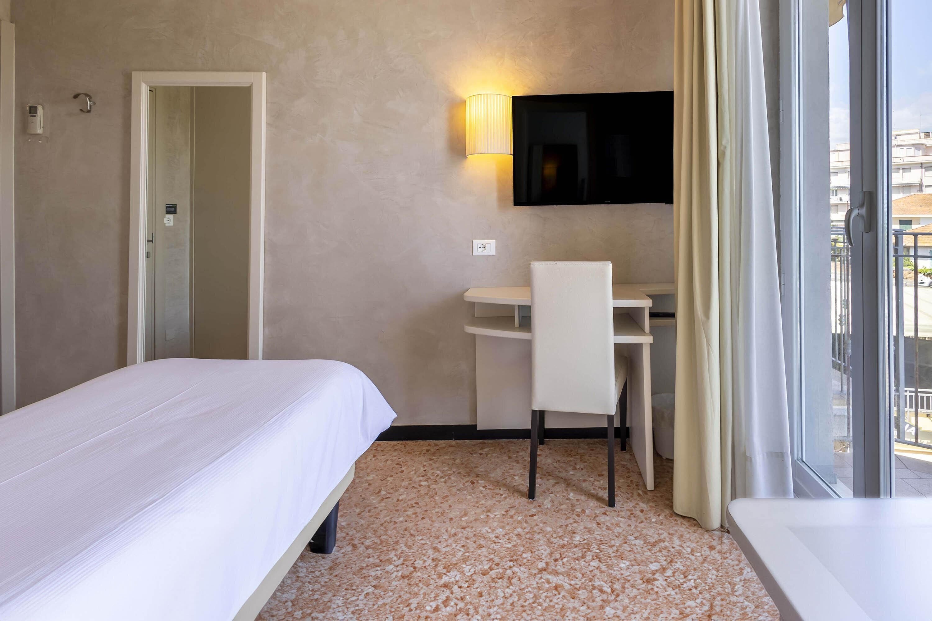Une chambre d’hôtel comprenant un lit, un bureau, une chaise, un téléviseur fixé au mur et une fenêtre avec des rideaux.