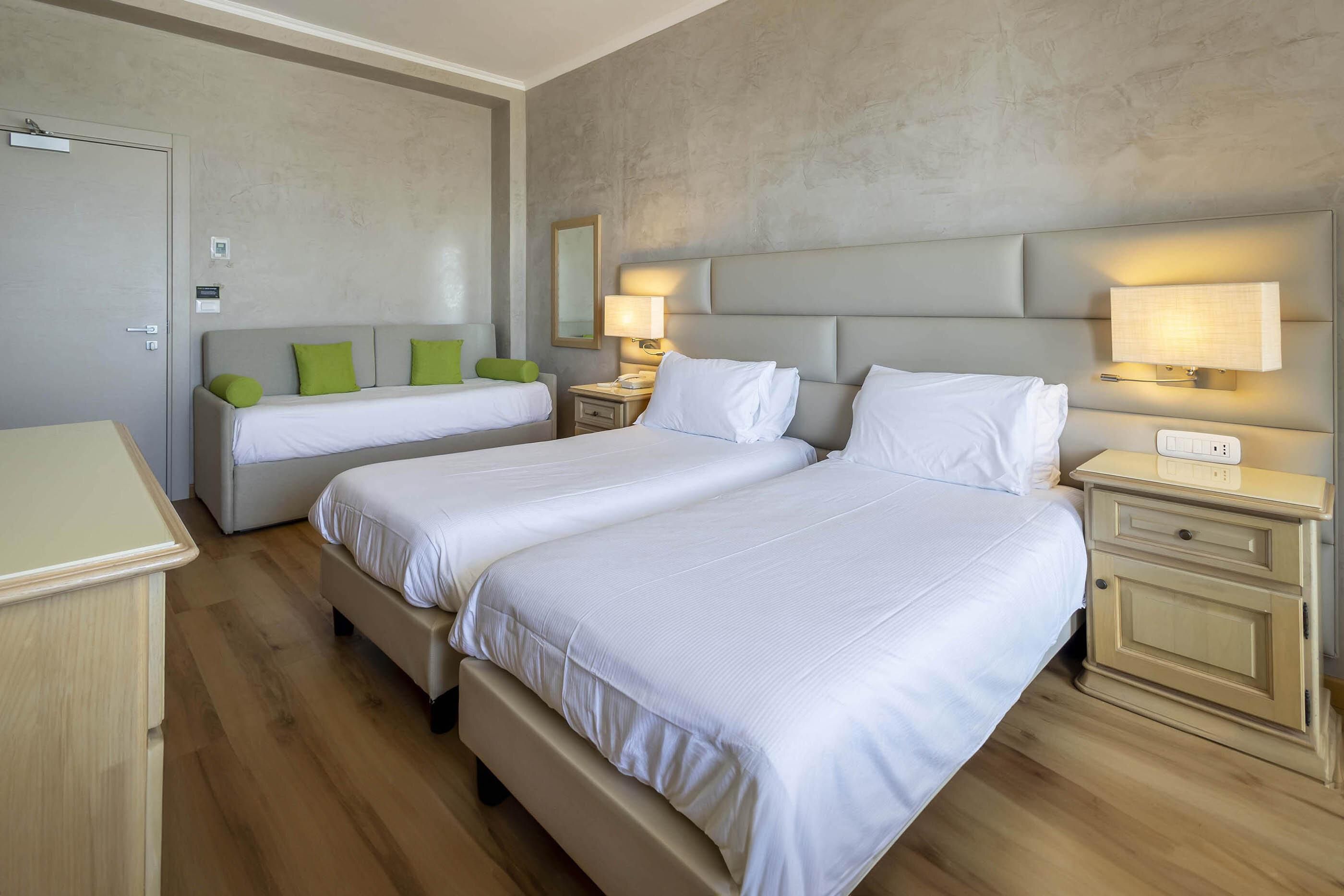 A hotel room with two beds, a wooden floor, a grey headboard, bedside lamps, and a small bedside table.