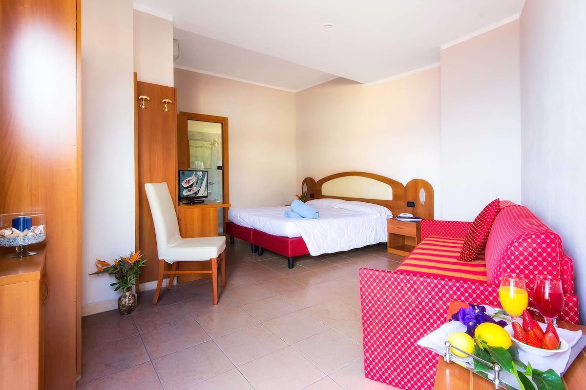 A hotel room with a bed, a desk, a chair, a sofa, and a breakfast tray with fruits and juice.
