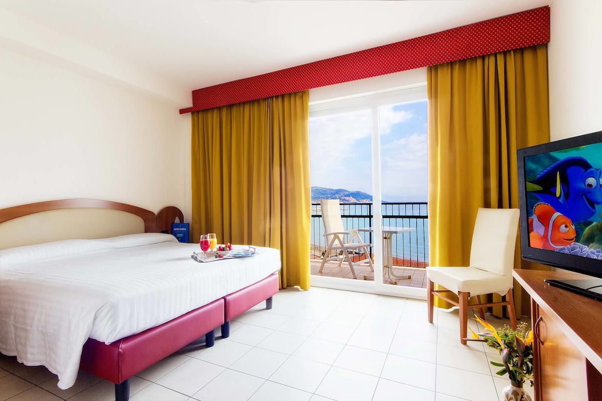 A hotel room with a bed, a TV, a balcony with yellow curtains, and a view of the sea.
