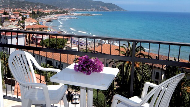 A balcony with white chairs and a small table, overlooking a beach with a clear view of the ocean and distant hills. - Piccolo Hotel (Diano Marina)