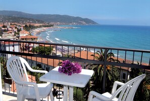 A balcony with white chairs and a small table, overlooking a beach with a clear view of the ocean and distant hills. - Piccolo Hotel (Diano Marina)