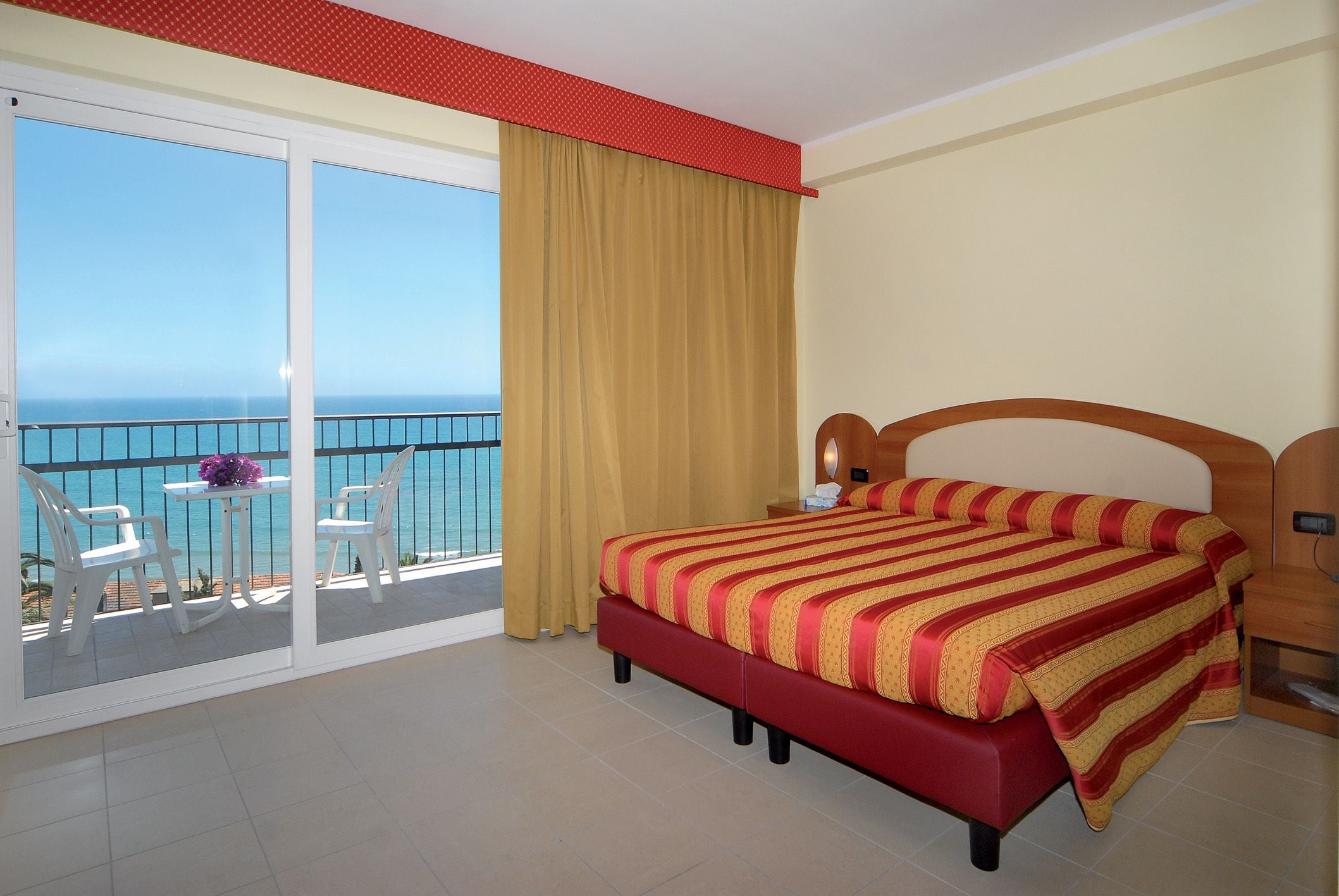Una habitación de hotel con una cama grande, un balcón con vistas al mar y una pequeña mesa con sillas.