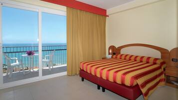 Una habitación de hotel con una cama grande, un balcón con vistas al mar y una pequeña mesa con sillas.