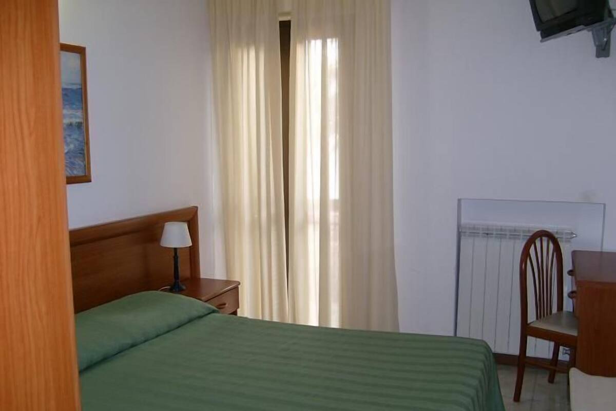 Una camera d'albergo con un letto, una scrivania, una sedia, un termosifone e una finestra con tende.
