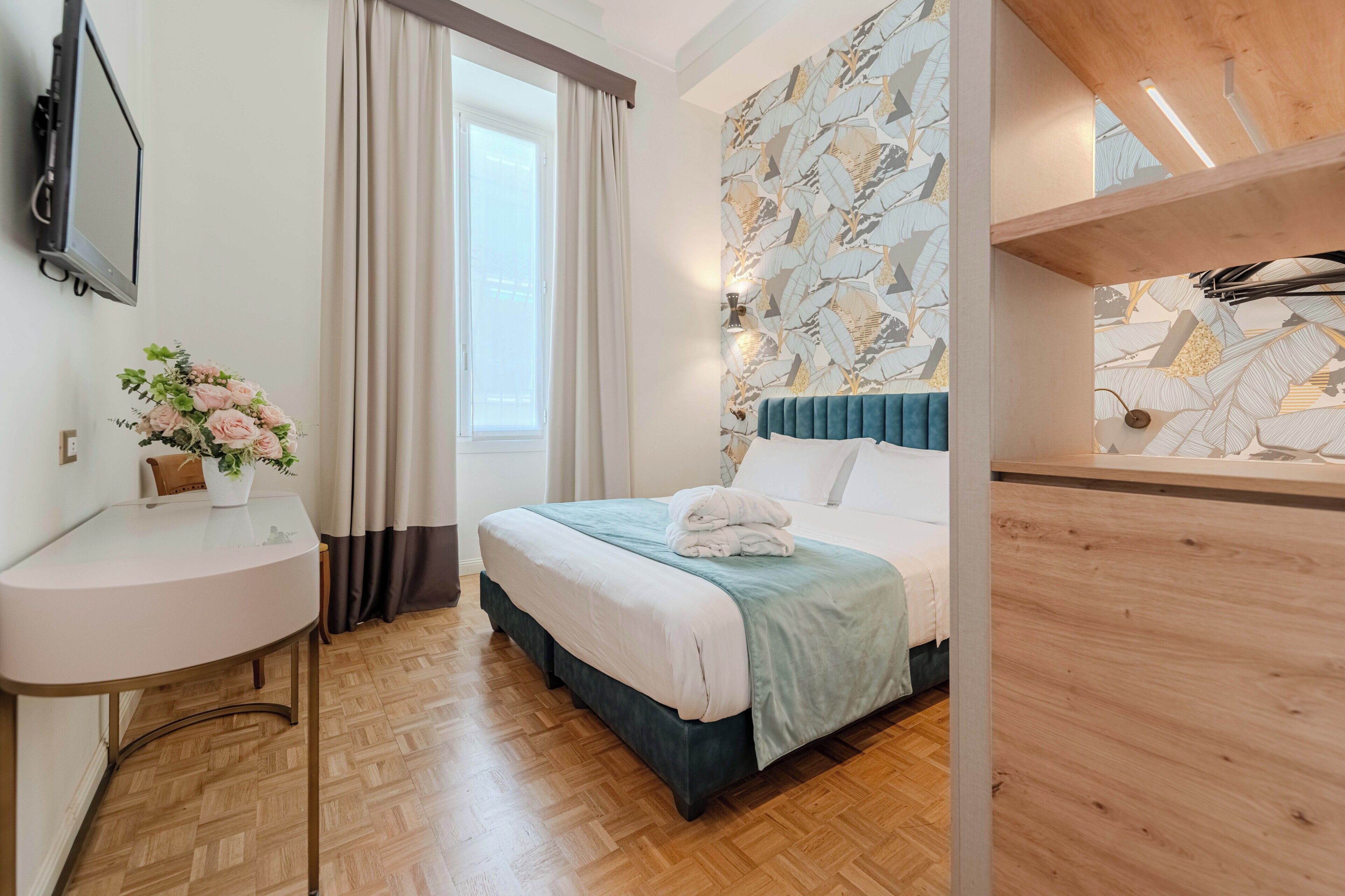 Ein Schlafzimmer mit Bett, Nachttisch, einem Fenster mit Vorhängen, einem an der Wand montierten Fernseher und einem Holzkopfende.