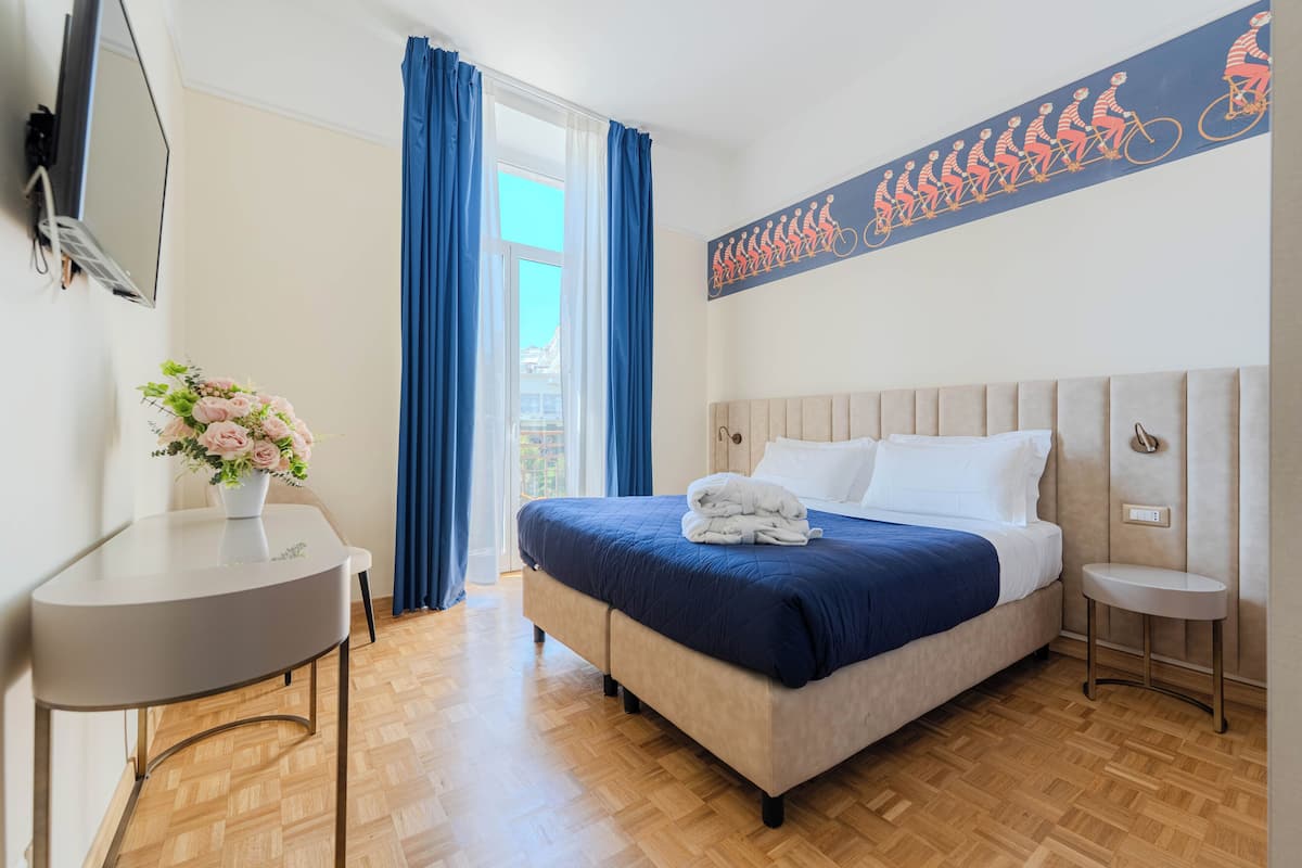 Una habitación de hotel con cama, una mesa, televisor y ventana con cortinas.