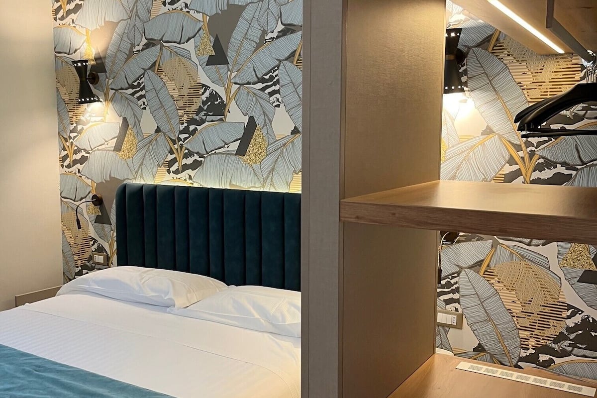 Habitación de hotel con cama, cabecera azul y pared con diseño de hojas. Se observa una estantería de madera con un piano encima.
