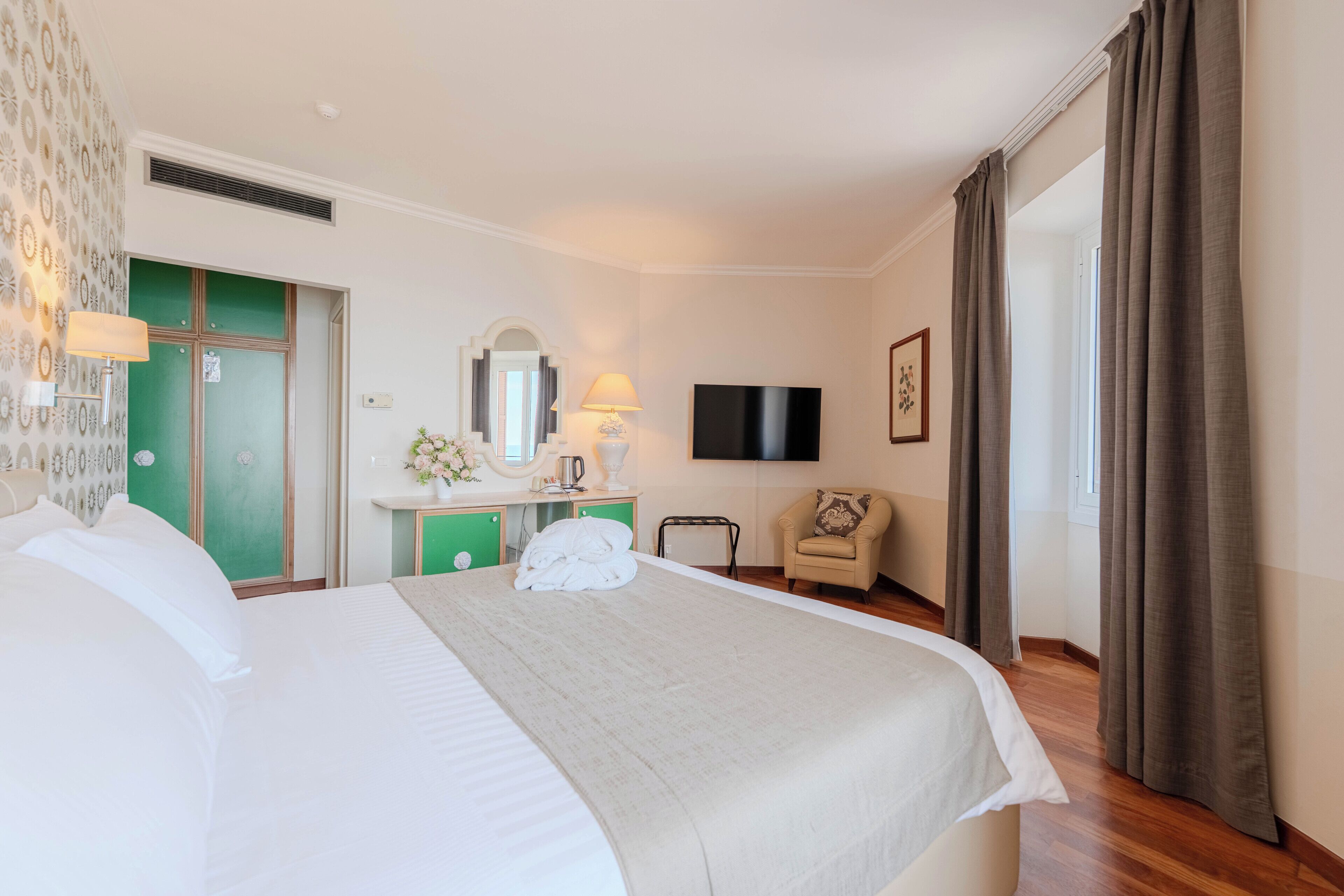 Una habitación de hotel con cama, tocador con espejo, televisión, silla y ventana con cortinas.