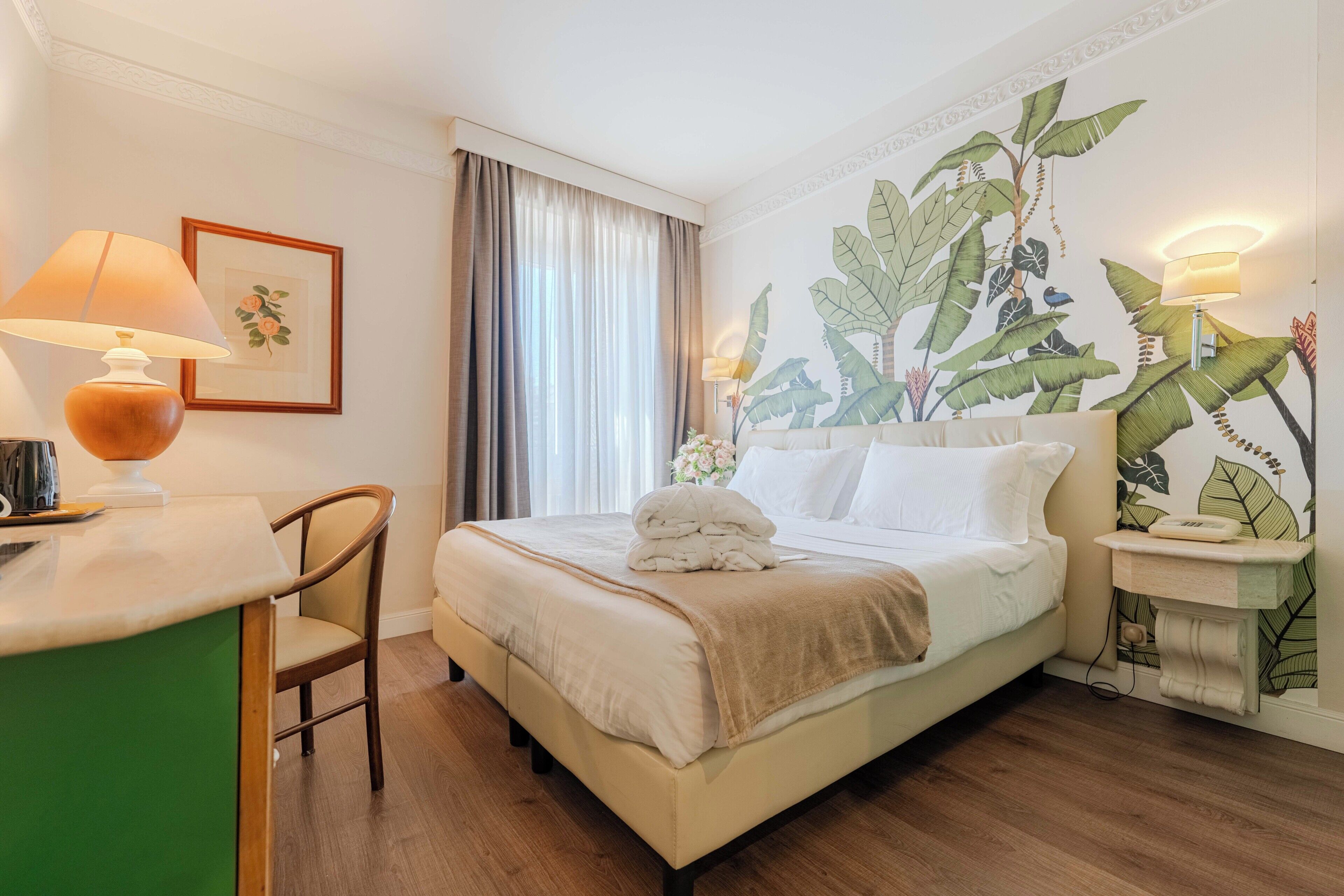 A hotel room with a bed, a desk, a chair, a lamp, a wall with a plant mural, and a window with curtains.