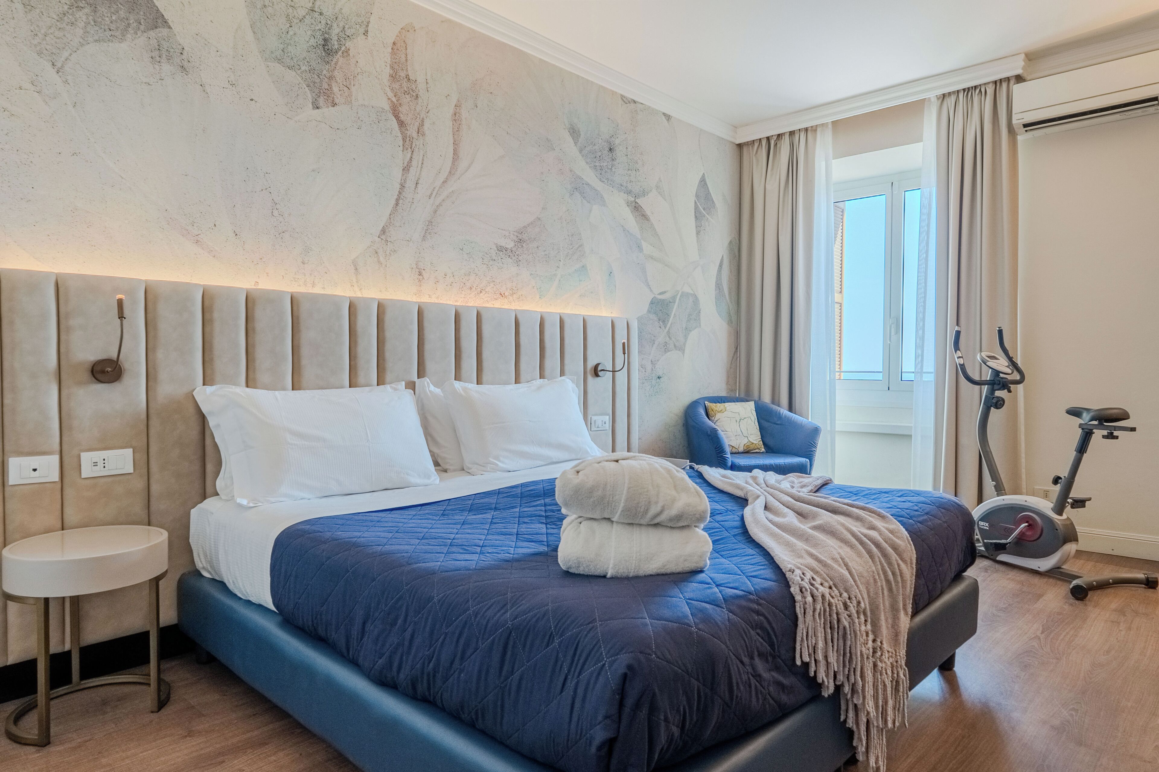 A modern bedroom with a large bed, a chair, an exercise bike, and a view of the outdoors.