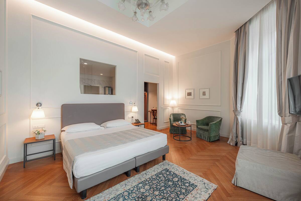 Una habitación de hotel moderna con una cama grande, mesitas de noche, un sillón verde, una pequeña mesa y un televisor.