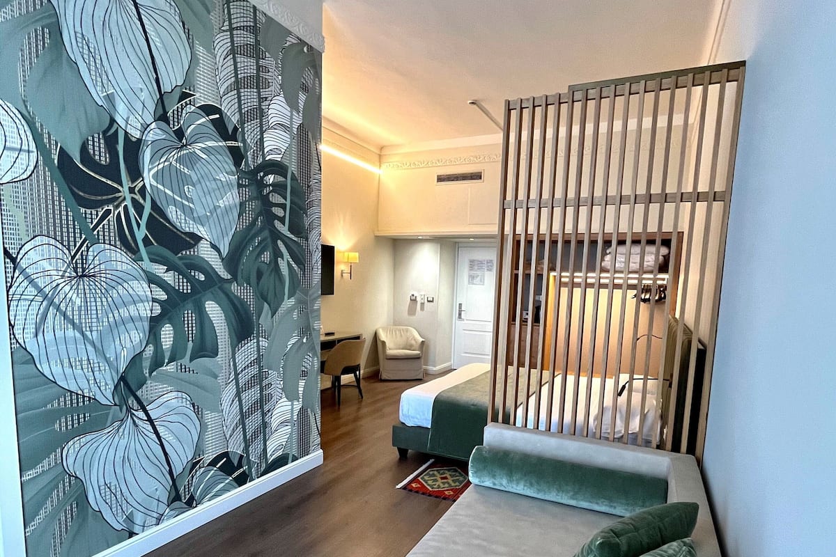 Una habitación de hotel moderna con un gran mural floral, un sofá con cojines verdes, una cama y una partición de madera.
