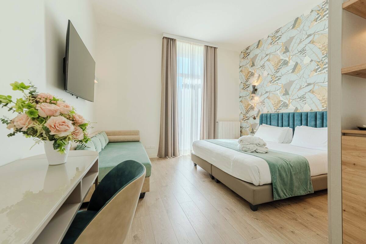Habitación de hotel con cama, escritorio con silla, televisor montado en la pared y ventana con cortinas.