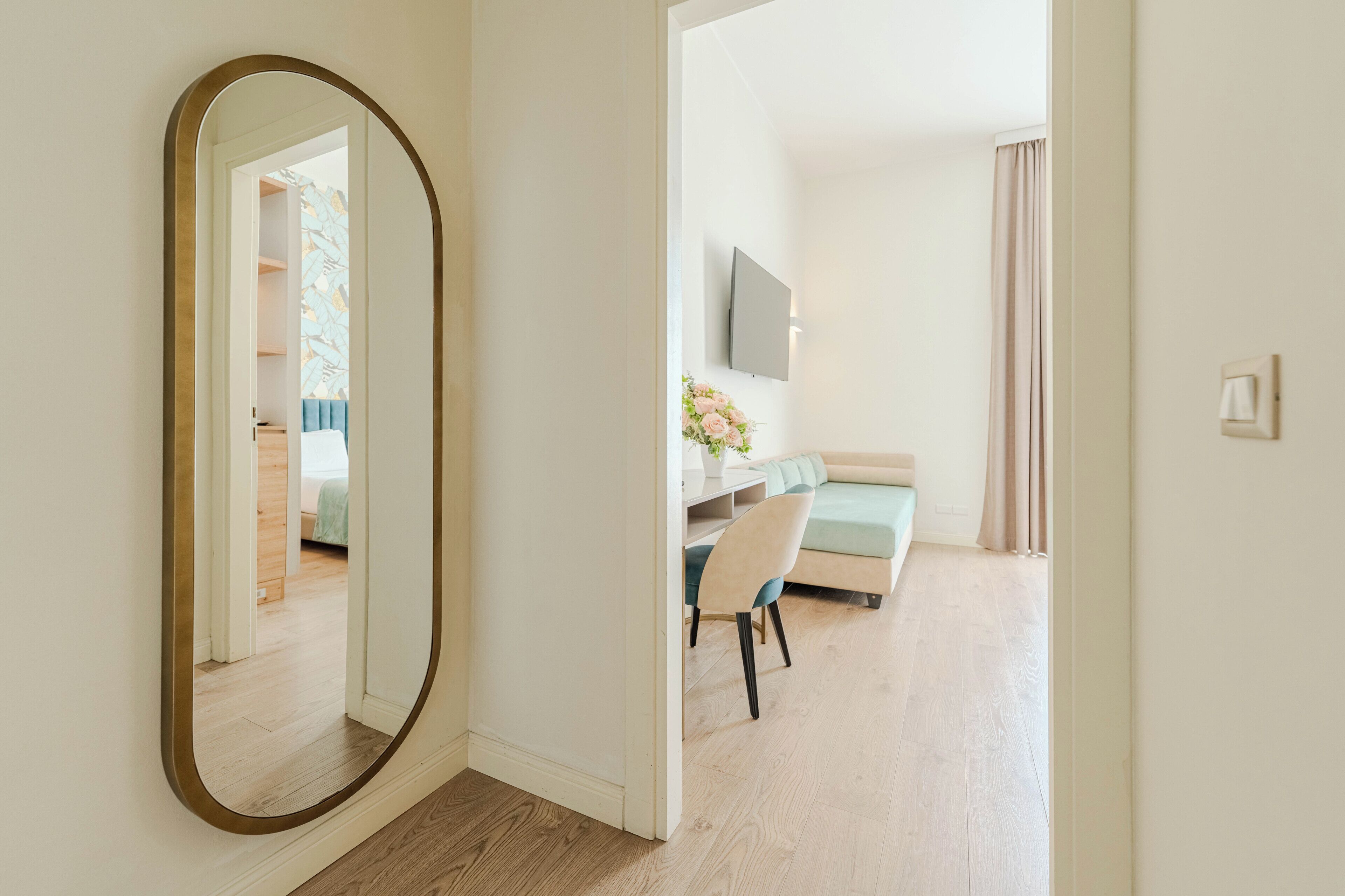 A room with a large mirror, a bed, a desk with a chair, a TV, and a window with curtains.