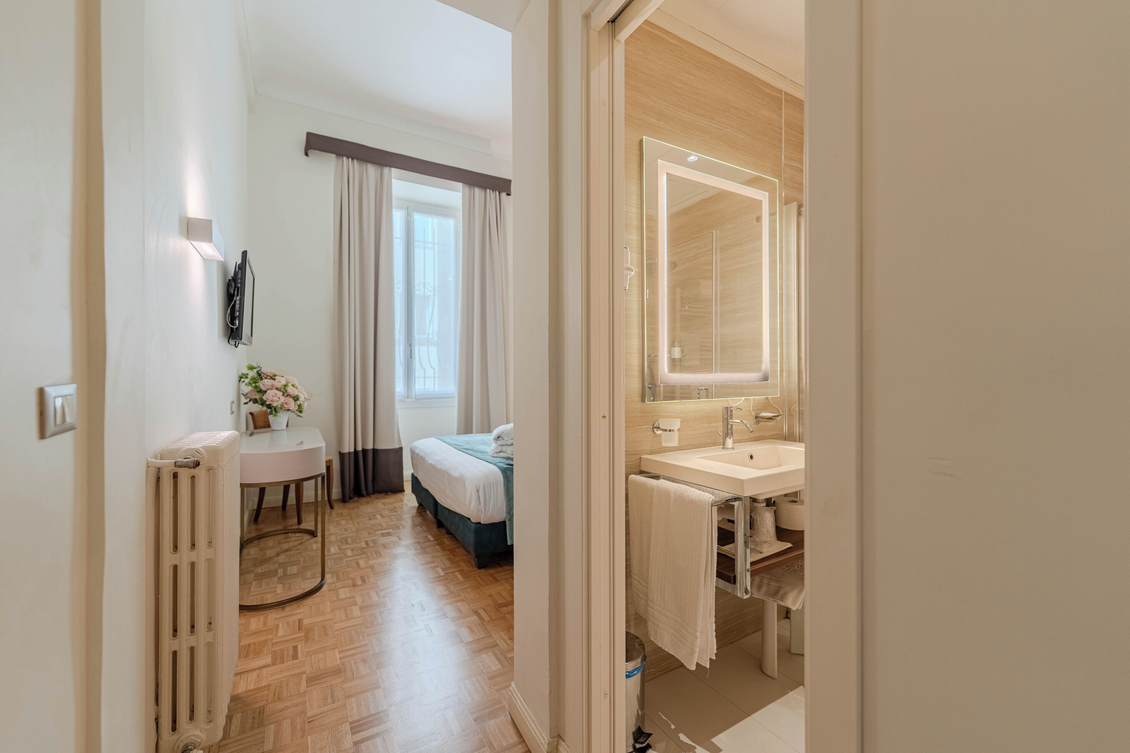 A hotel room with a bed, a round table with a vase, a radiator, a bathroom with a mirror and sink.