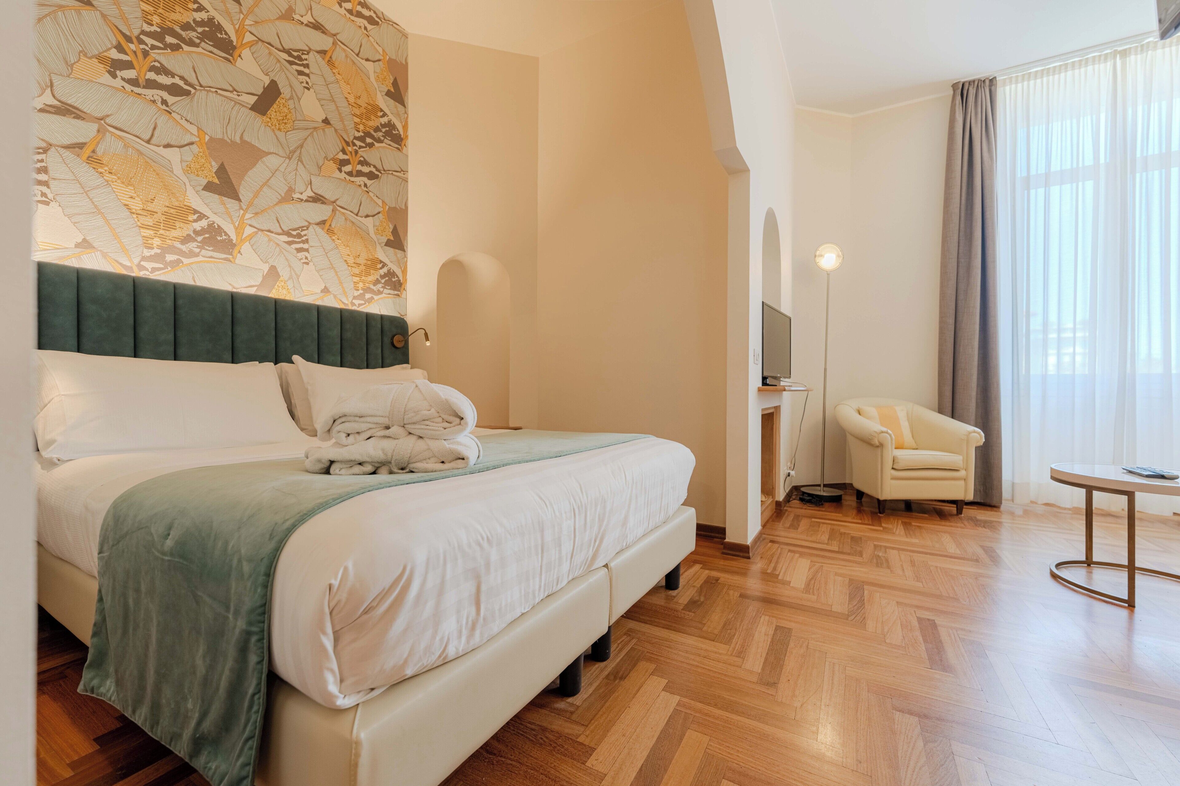 Habitación de hotel con cama, una silla, una mesa, televisor y un ventanal con cortinas.