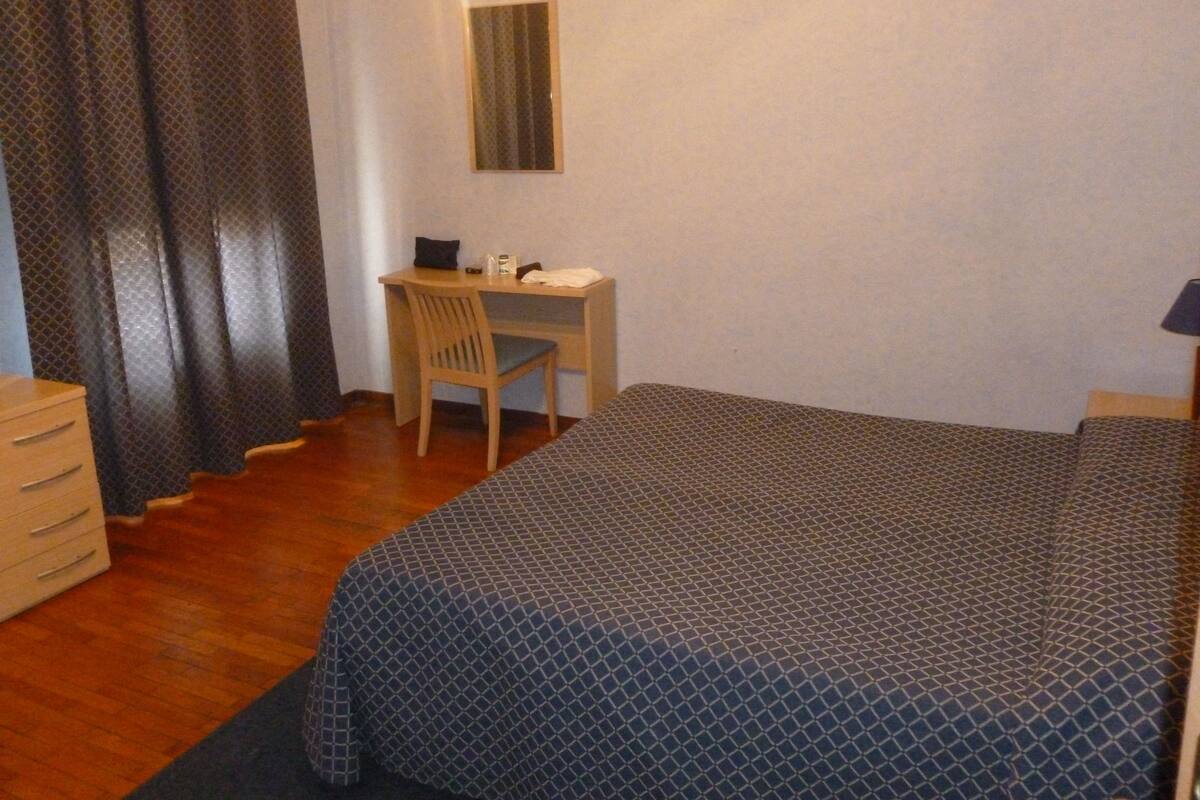 A bedroom with a bed, a desk, a chair, a dresser, and a window with curtains.