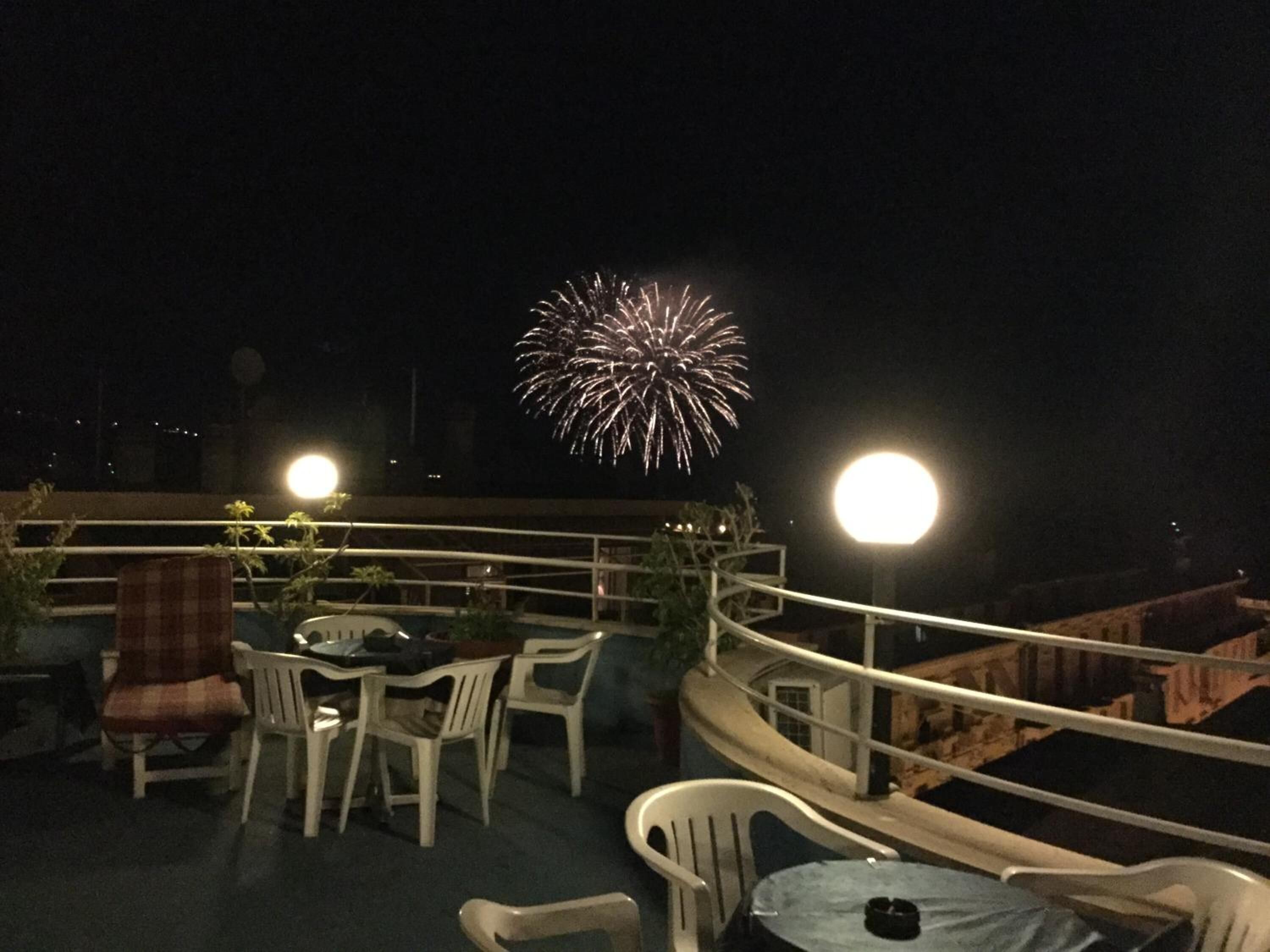 Una terraza en la azotea con sillas y mesas de plástico blanco, barandilla y vista de fuegos artificiales en el cielo nocturno.