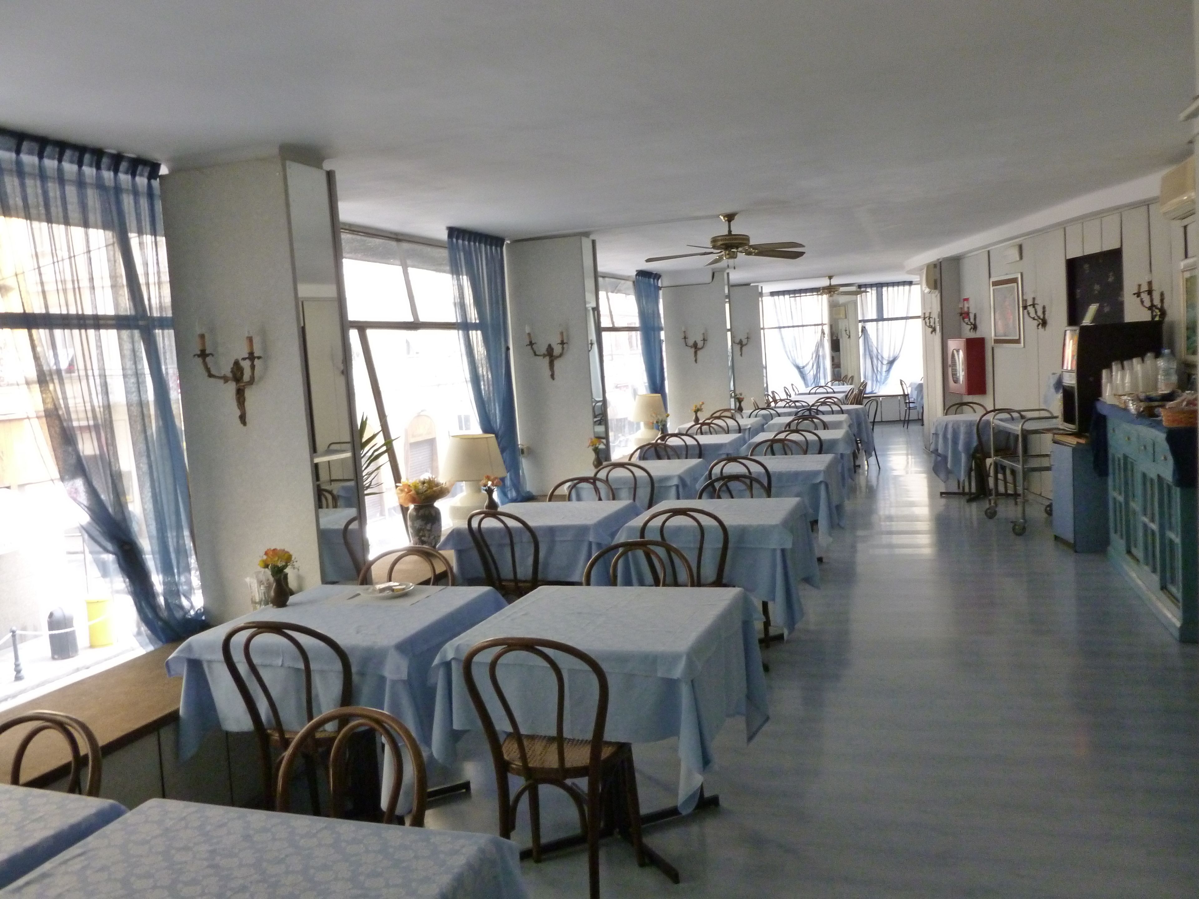 A dining area with multiple tables and chairs, large windows with blue curtains, and ceiling fans.