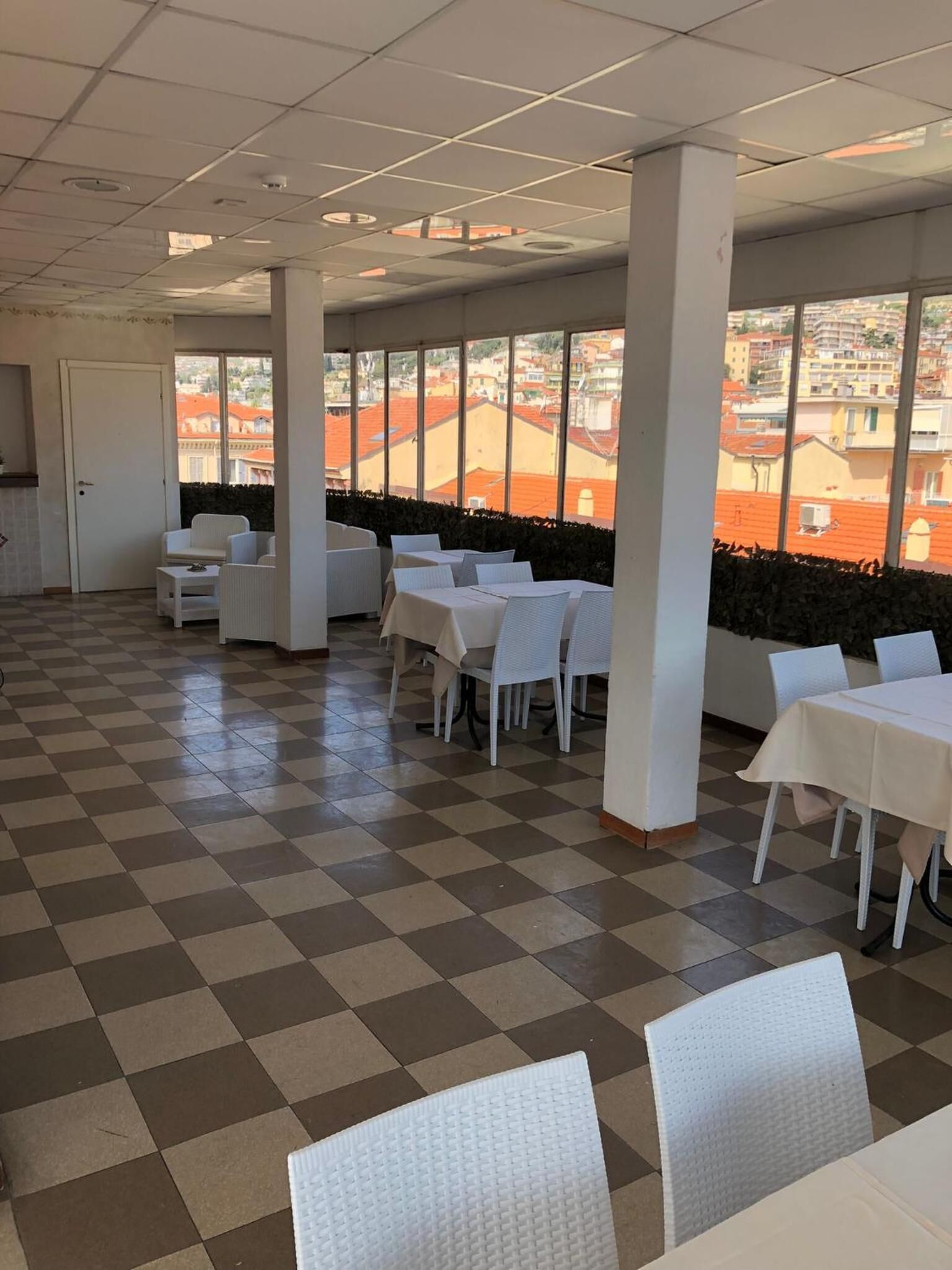 A spacious room with checkered flooring, white tables and chairs, large windows with a view of buildings, and a door.