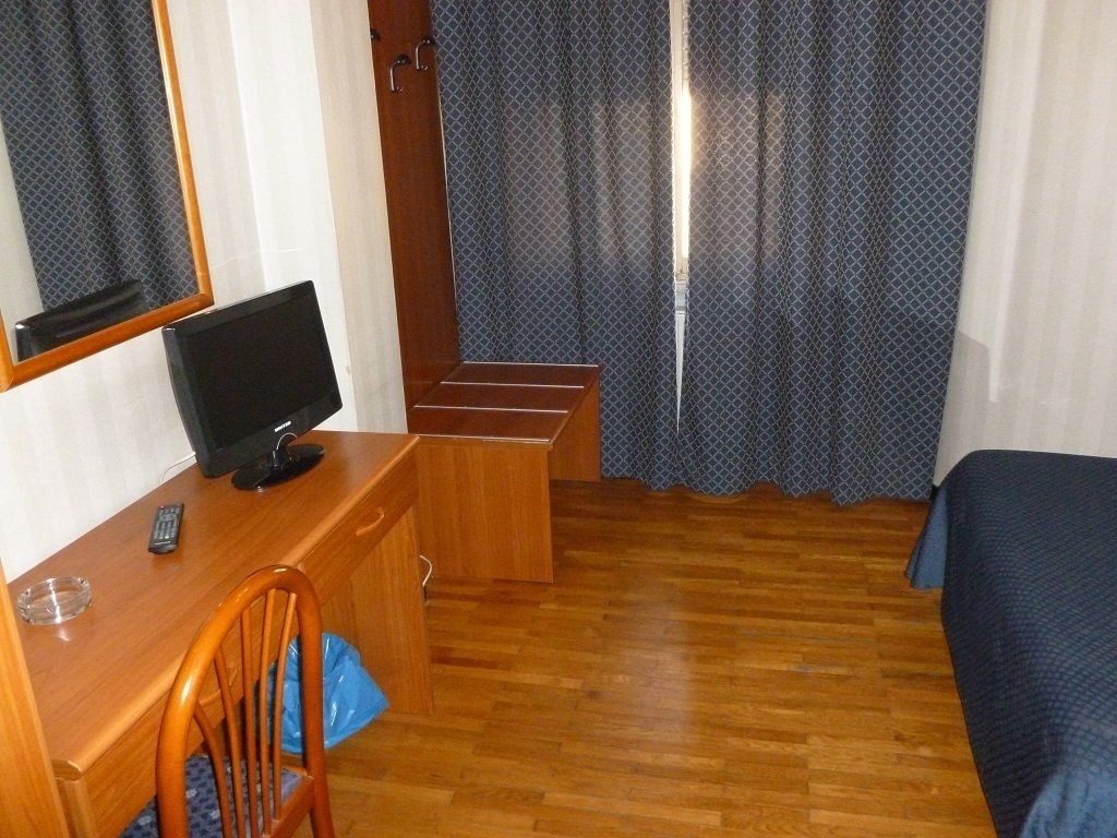 Una habitación de hotel con un escritorio de madera, televisión, una silla, cama y ventana con cortinas.