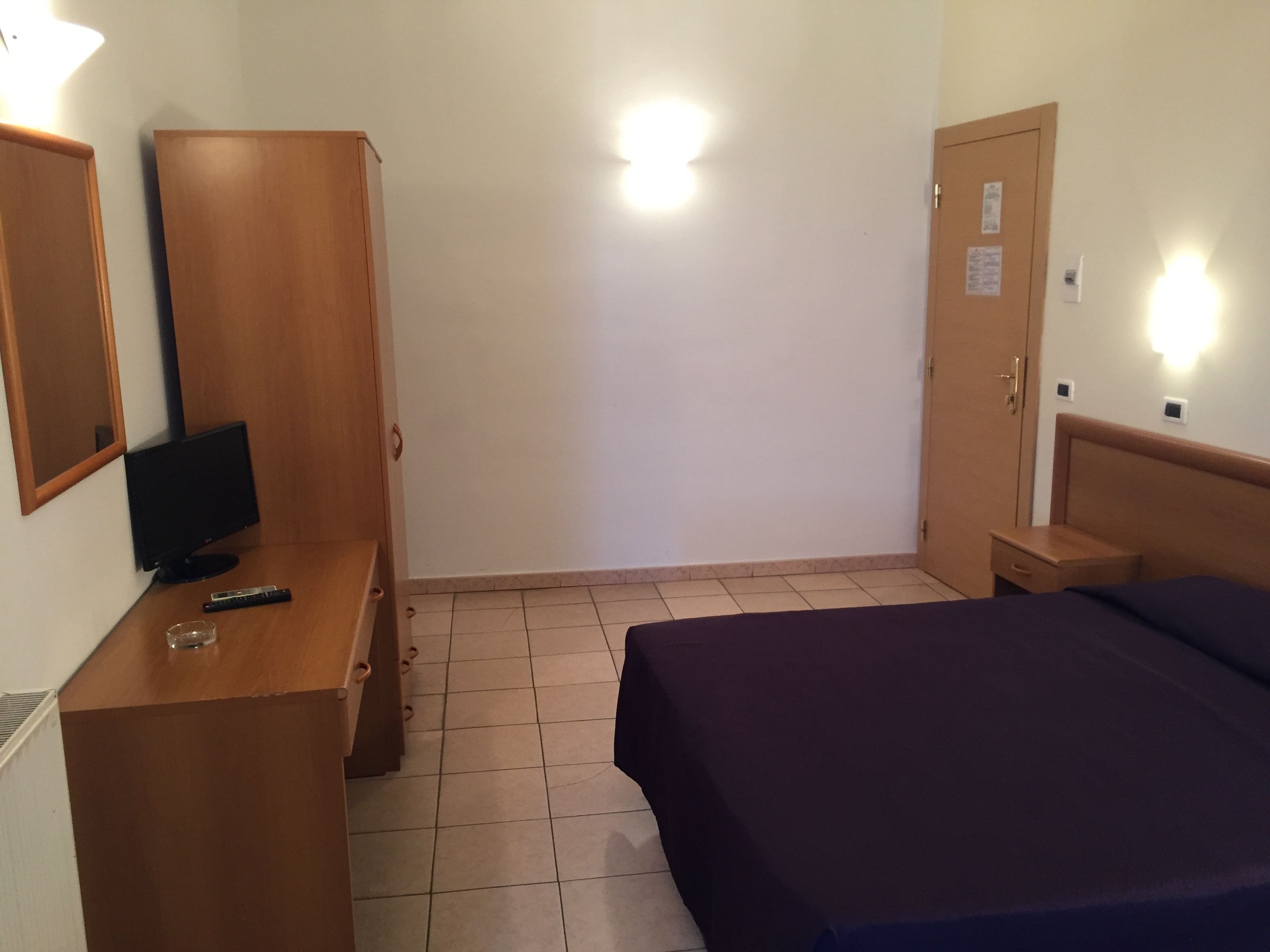 A hotel room with a bed, a desk with a computer and a remote control, a wardrobe, and a door with a light switch.